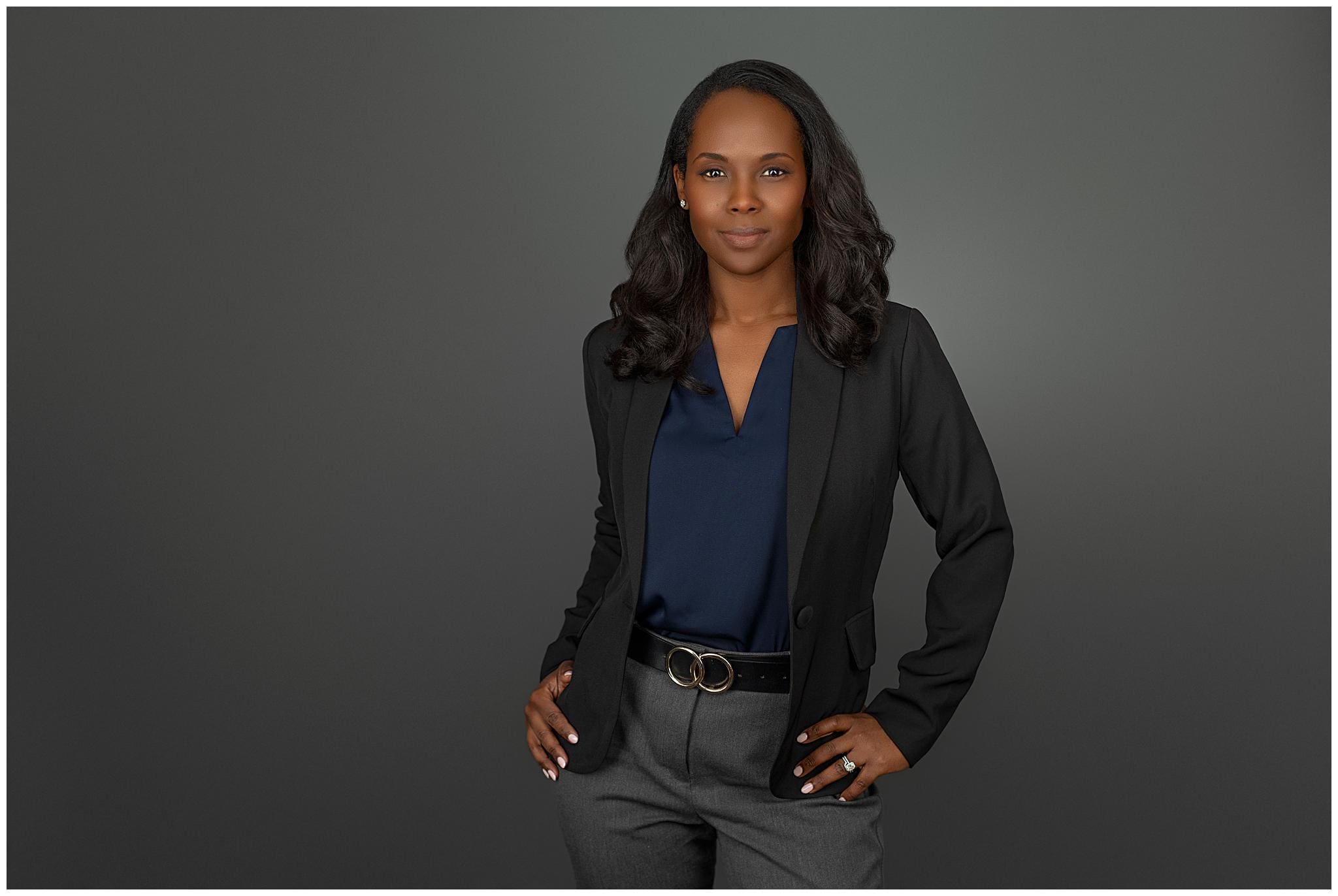 A confident woman with long dark hair wears a navy blouse, dark blazer, and gray pants. Posing like a natural in headshot photos, her hands rest on her hips as she stands against a plain gray background, smiling slightly.
