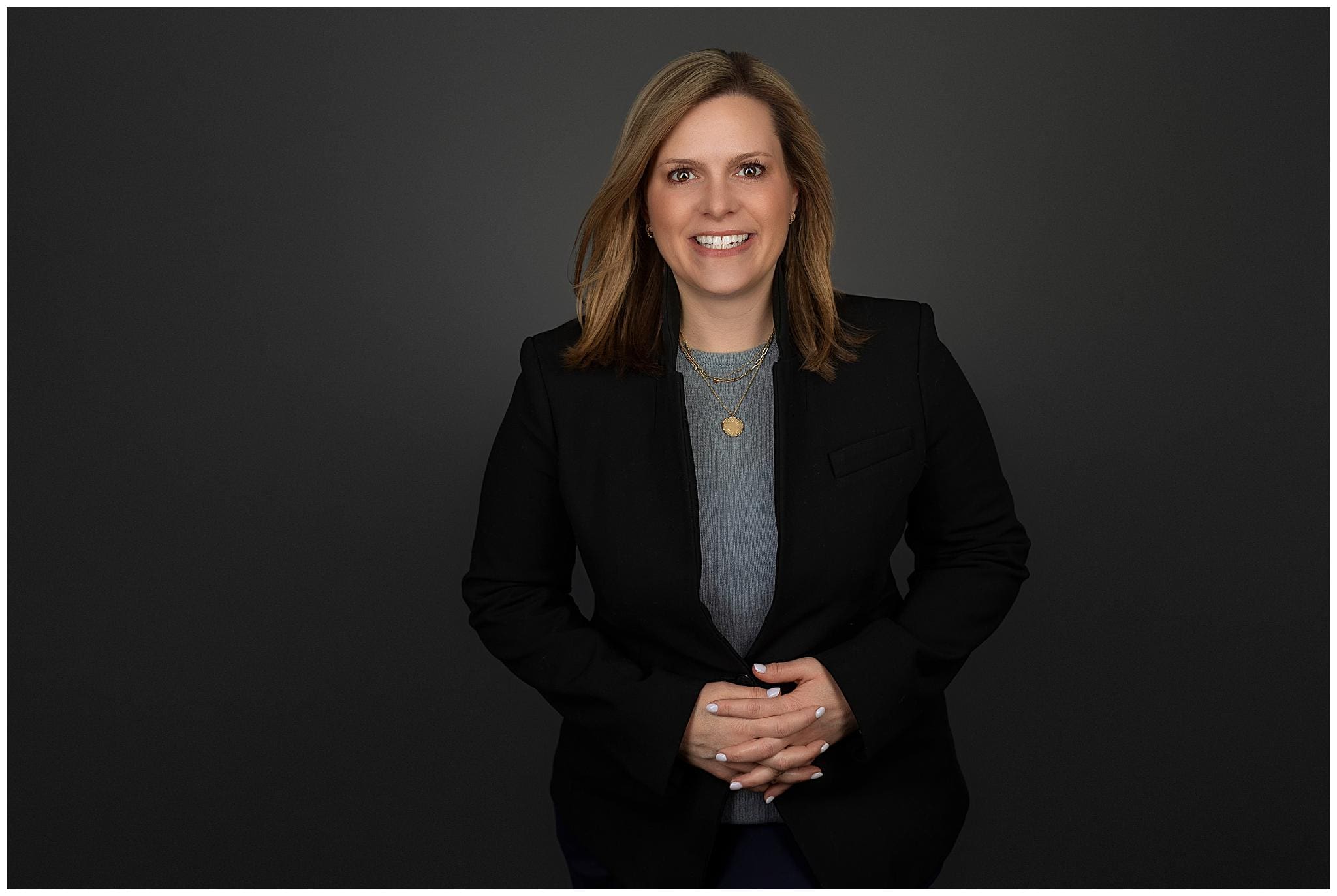 A woman with shoulder-length brown hair, wearing a black blazer over a gray top, smiles at the camera in this professional headshot. She is standing against a dark gray background with her hands clasped in front of her.
