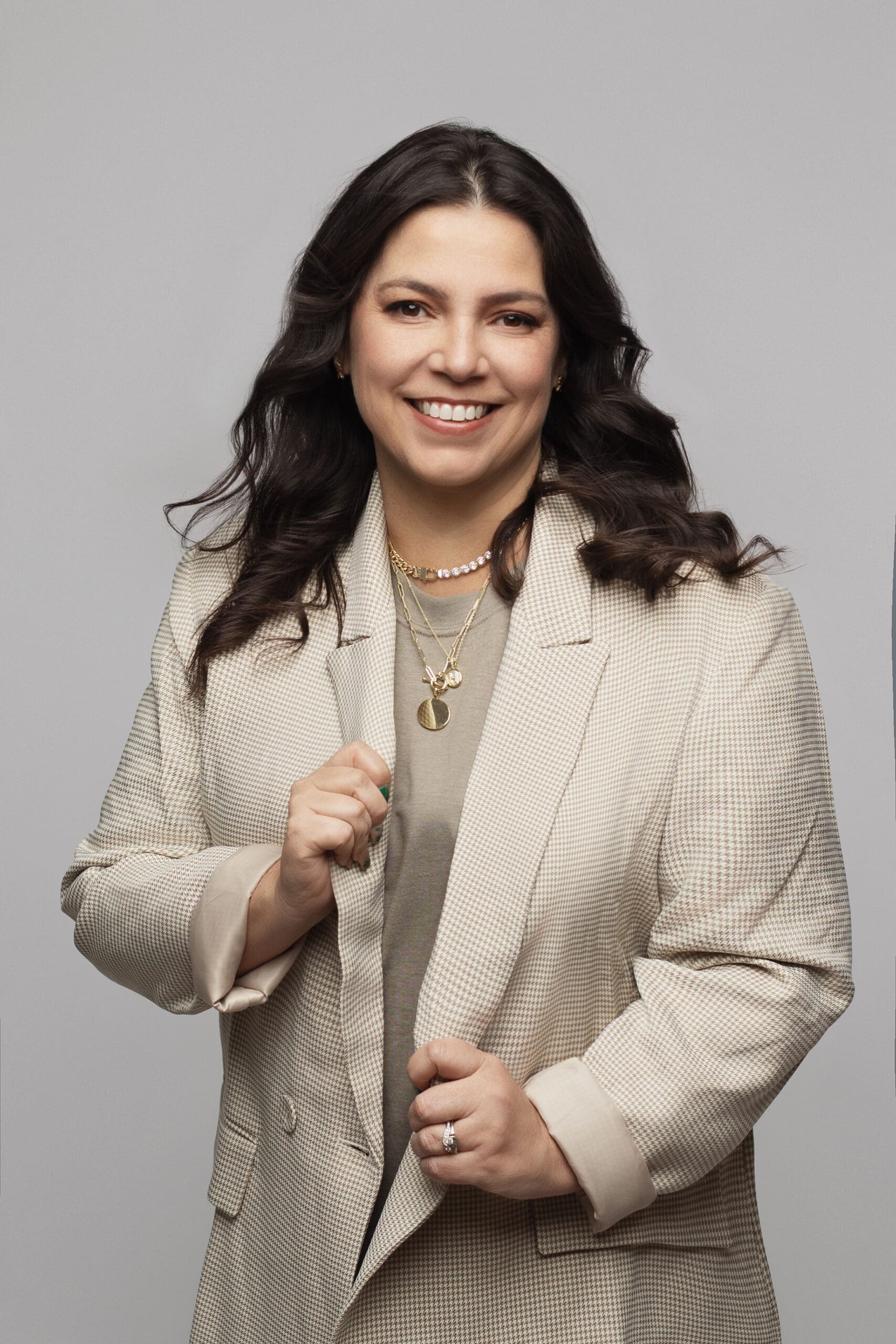 Smiling person with long dark hair wearing a beige blazer, taupe shirt, and layered necklaces against a plain gray background during her Connecticut Branding session.