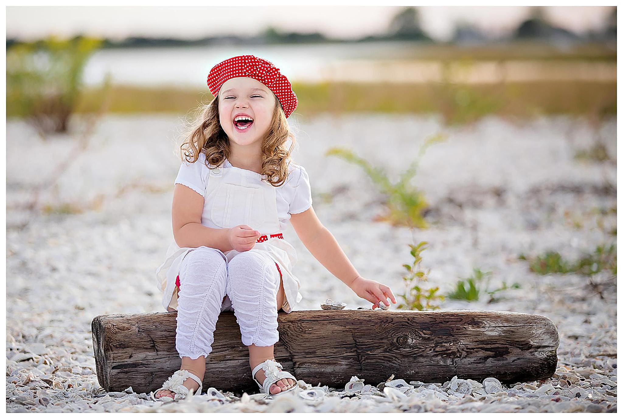 A young girl with curly hair, wearing a red patterned beret and white outfit, sits on a weathered log at a pebble-covered beach. She is smiling brightly and holding a small shell. The background features blurred natural scenery on a Milford Beach.