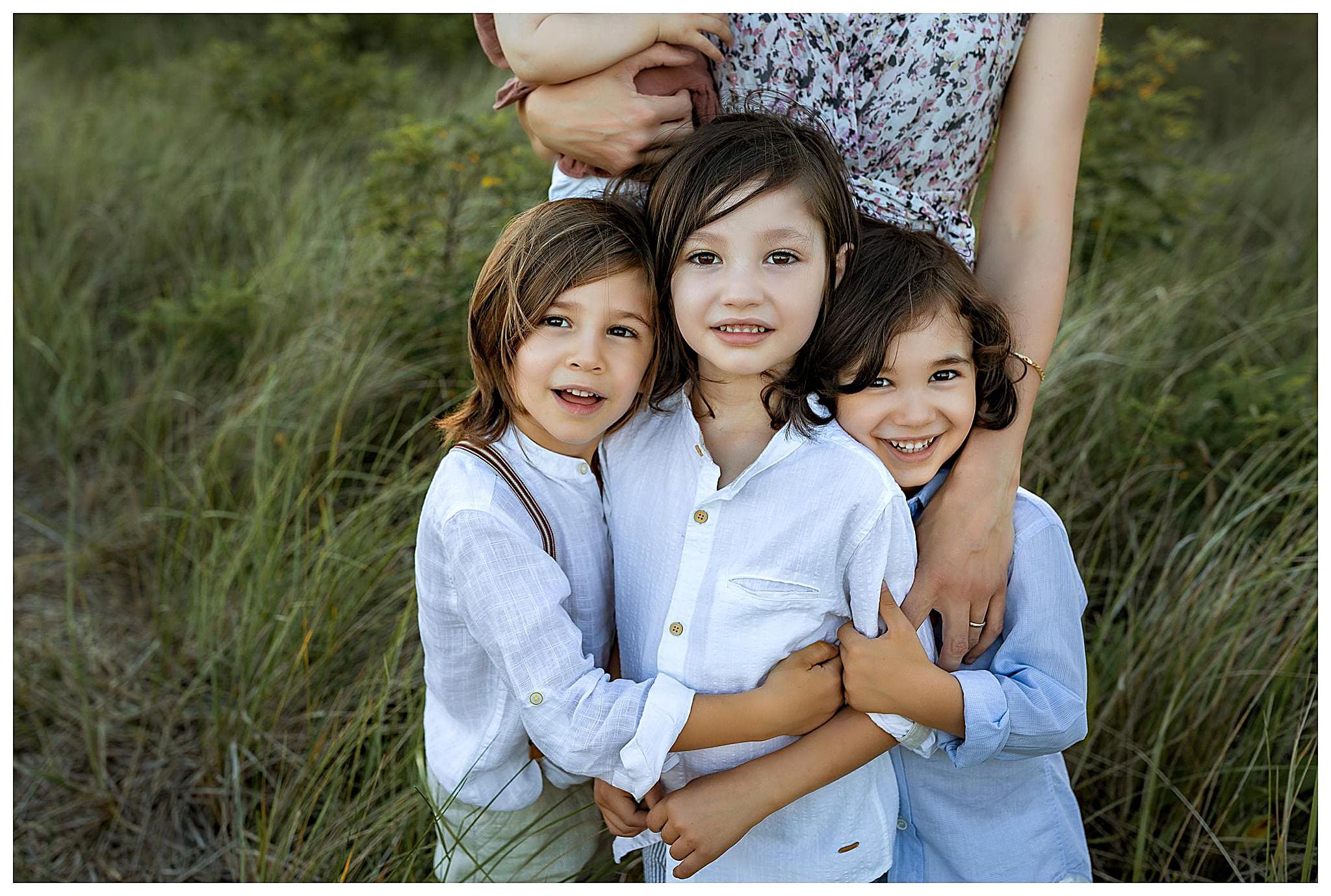 Three children with long hair and light-colored shirts stand closely together, smiling. An adult partly visible behind them gently holds them. The background features greenery and tall grass on a beach in Connecticut.
