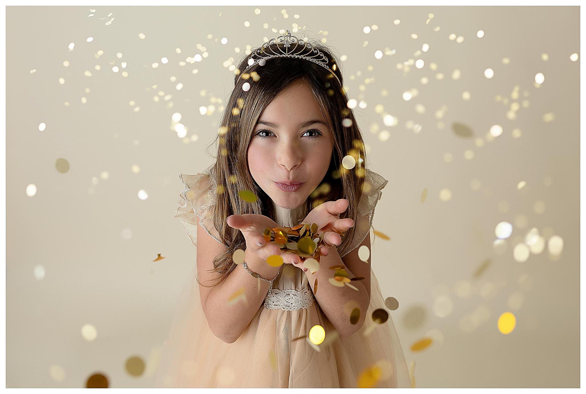 Young girl wearing a tiara and a light-colored dress blows a handful of gold confetti towards the camera. The background is a soft, neutral color with sparkling bokeh effects, creating a magical, celebratory atmosphere.