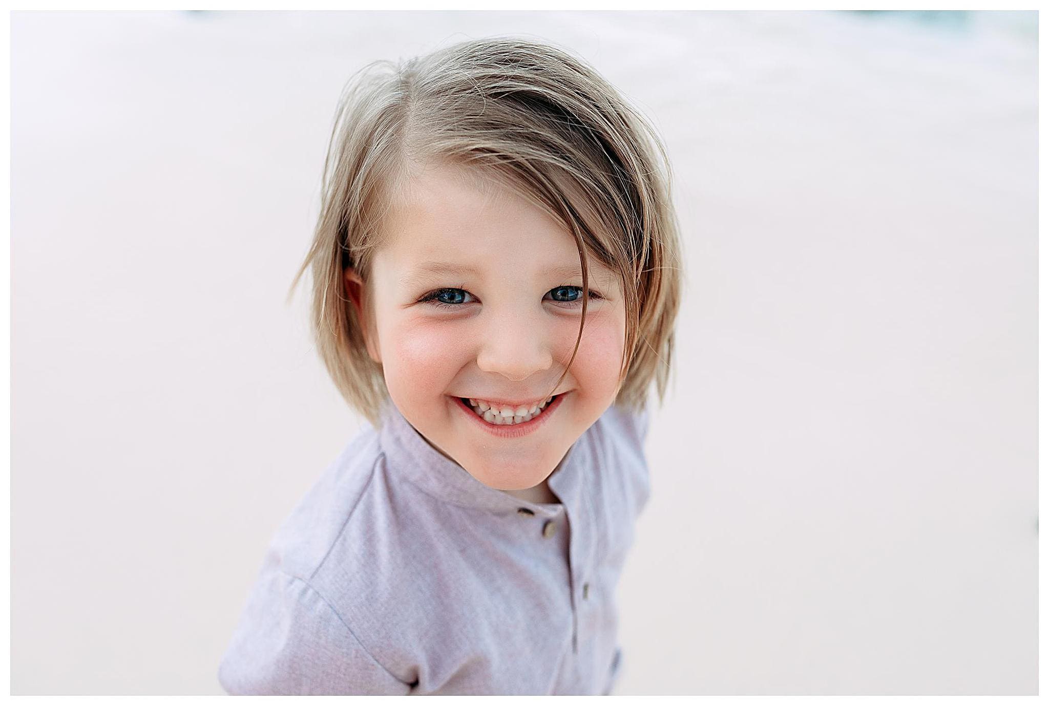 A smiling child with shoulder-length blond hair stands on a sandy beach, wearing a light gray button-up shirt. The background is a blurred, light beige sand.