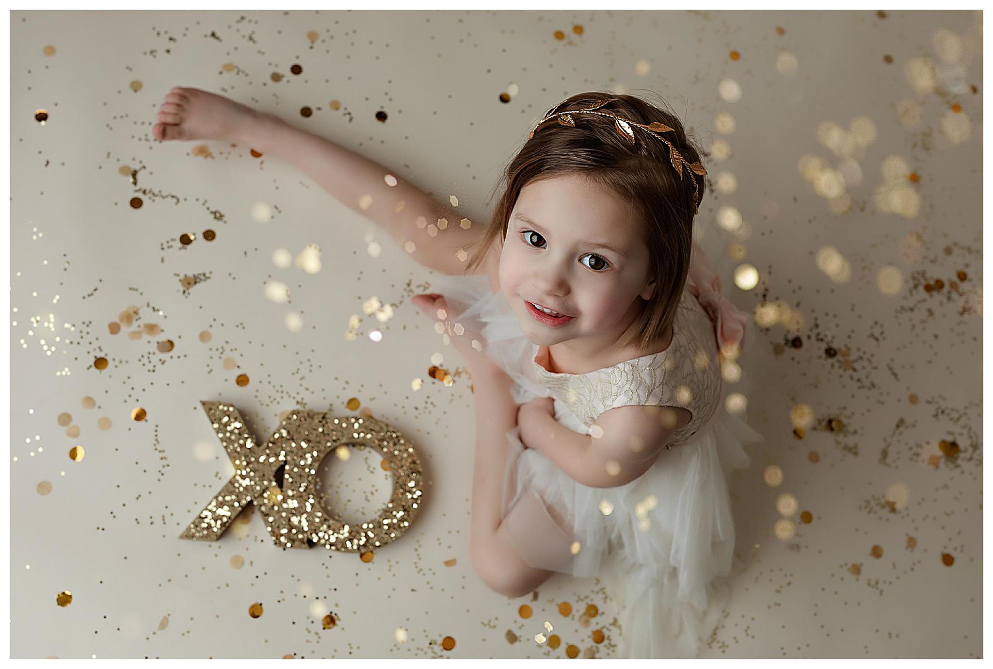 A young girl in a white dress looks upwards, smiling, surrounded by gold glitter and confetti. She wears a headband and is seated near glittery XO letters on the floor.