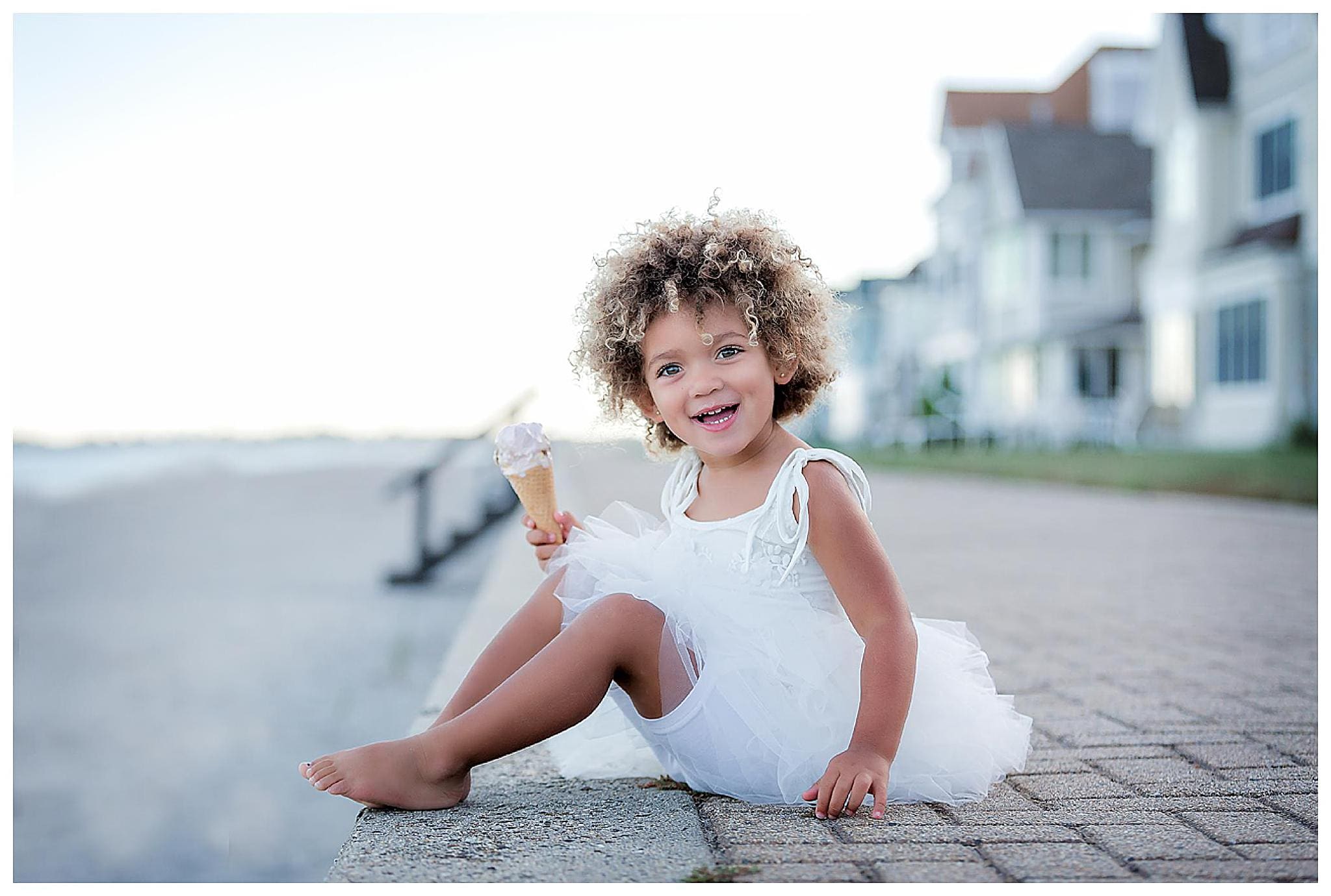 A young child with curly hair, wearing a white dress, sits on a stone ledge by the beach, smiling and holding an ice cream cone. The background features blurred seaside houses and a calm sea.