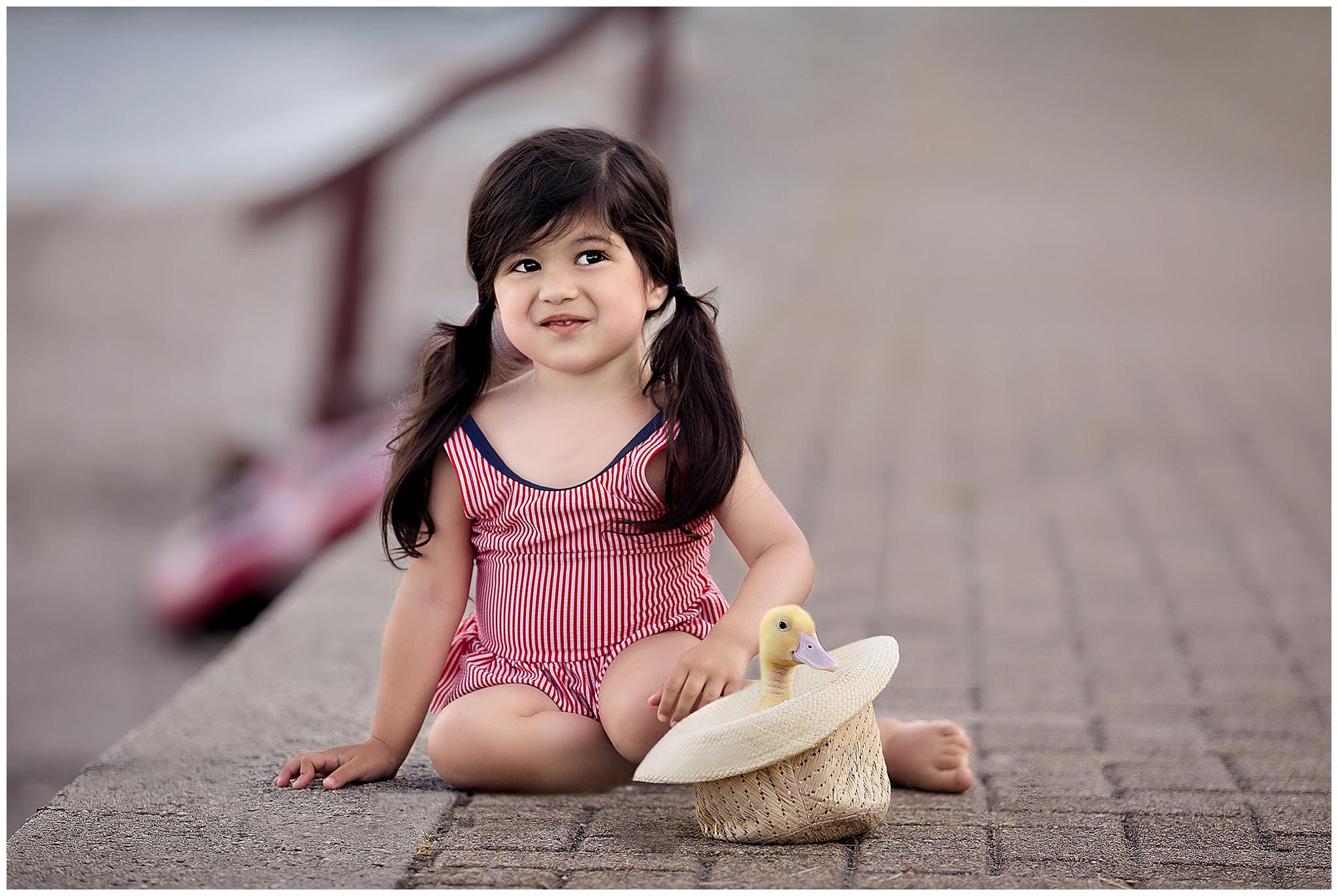 A young girl sits on a stone path wearing a red and white striped outfit. She has dark hair in pigtails and is smiling. Next to her is a straw hat containing a yellow rubber duck. The background is blurred.