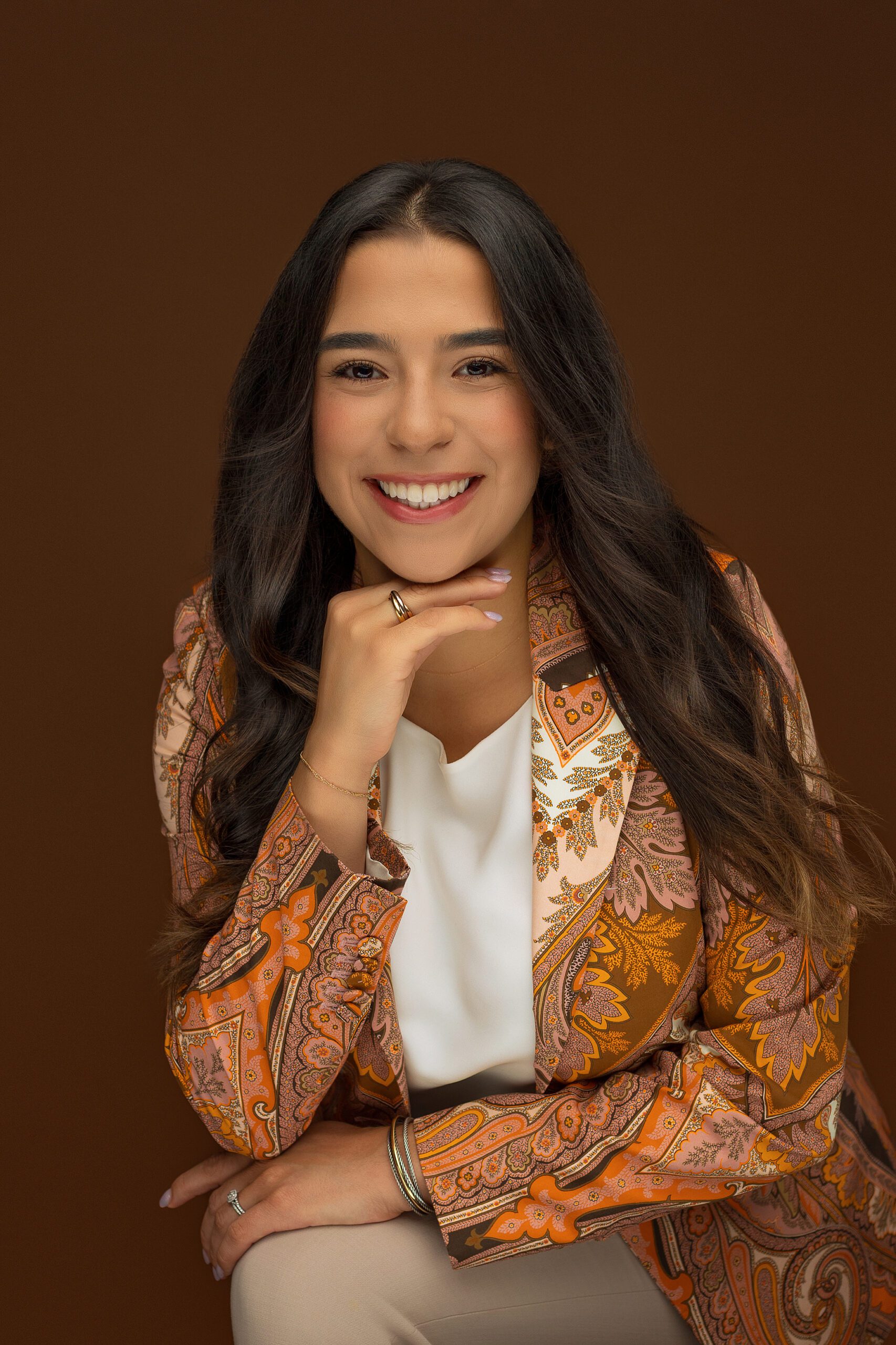 A woman with long dark hair smiles warmly at the camera. She is wearing a patterned orange blazer over a white blouse and is seated with her hand resting on her chin, against a brown background during her CT headshot session.