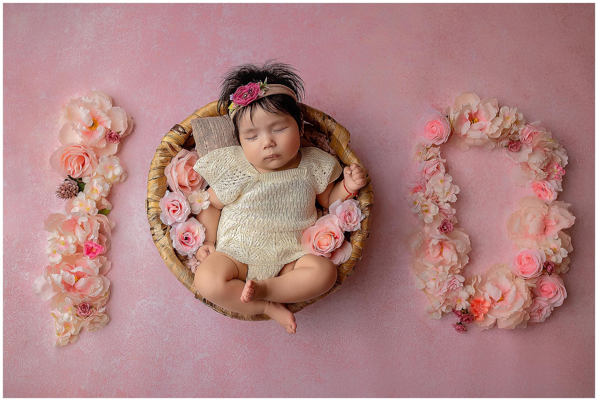 A baby sleeps in a round basket adorned with pink and white flowers, wearing a cream outfit and floral headband. The number 100 is formed by flowers on a soft pink background celebrating her milestone