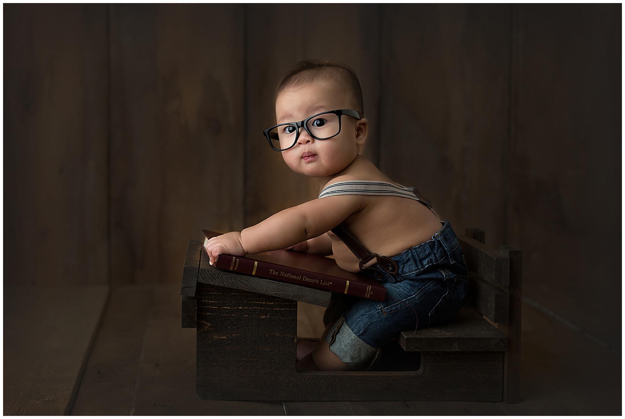 A baby, captured in a delightful sitter session, wears large black glasses and denim overalls while sitting in a wooden crate, holding a book titled The National Dream Lamp. The dark wooden background enhances the vintage look of this charming scene.