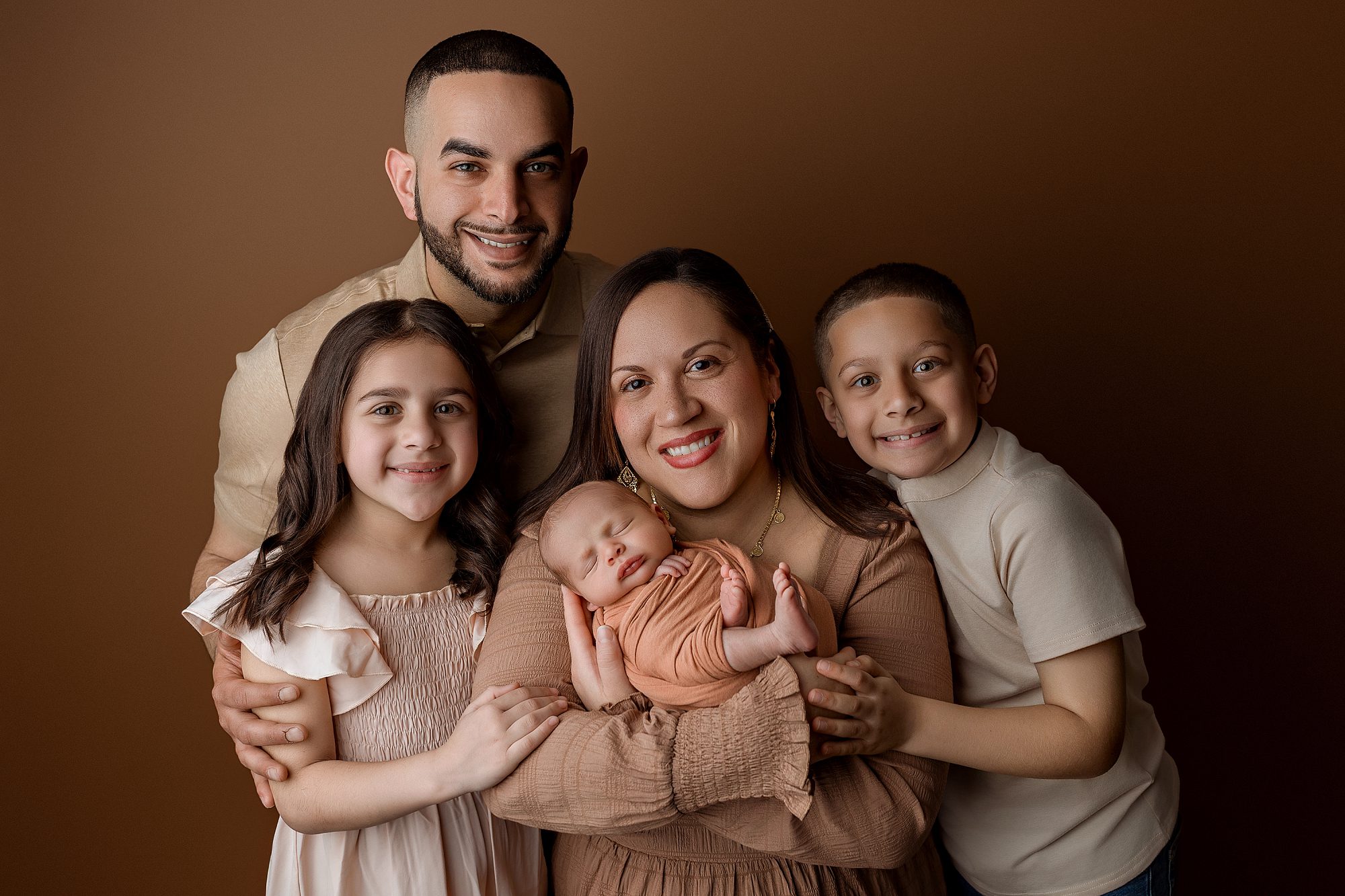 A smiling family of five poses closely together against a brown background. Parents stand behind their two children, while the mother cradles a newborn baby wrapped in a peach blanket. Everyone is dressed in neutral tones for their connecticut newborn photo session.