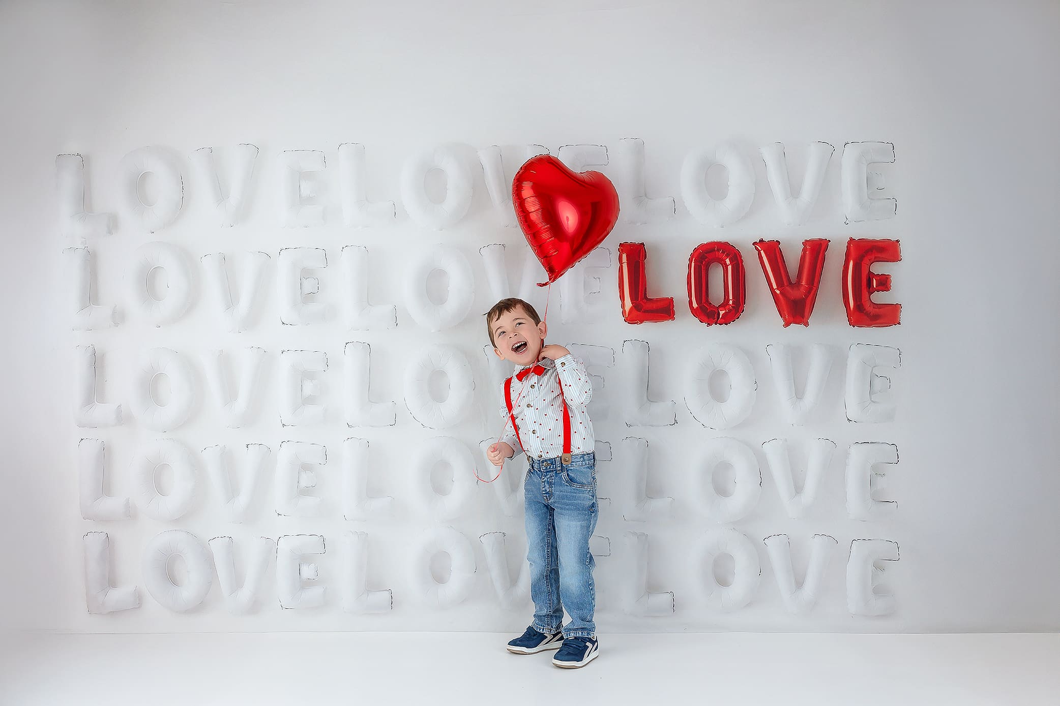A smiling child wearing jeans, a white shirt with red suspenders, and a bow tie holds a red heart-shaped balloon. The background features repeated white and red LOVE balloon letters on a light wall.