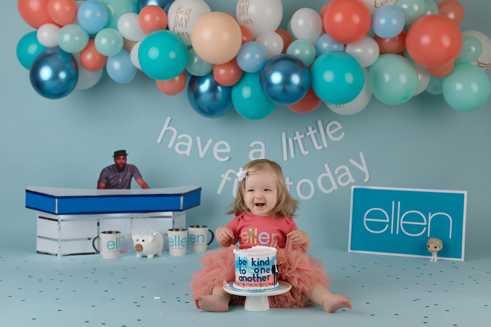 A smiling toddler in a pink tutu sits on the floor with a blue cake that says be kind to one another. Behind her are colorful balloons and ellen signs, with a light blue backdrop and playful decorations mimicking the Ellen DeGeneres Show setup.