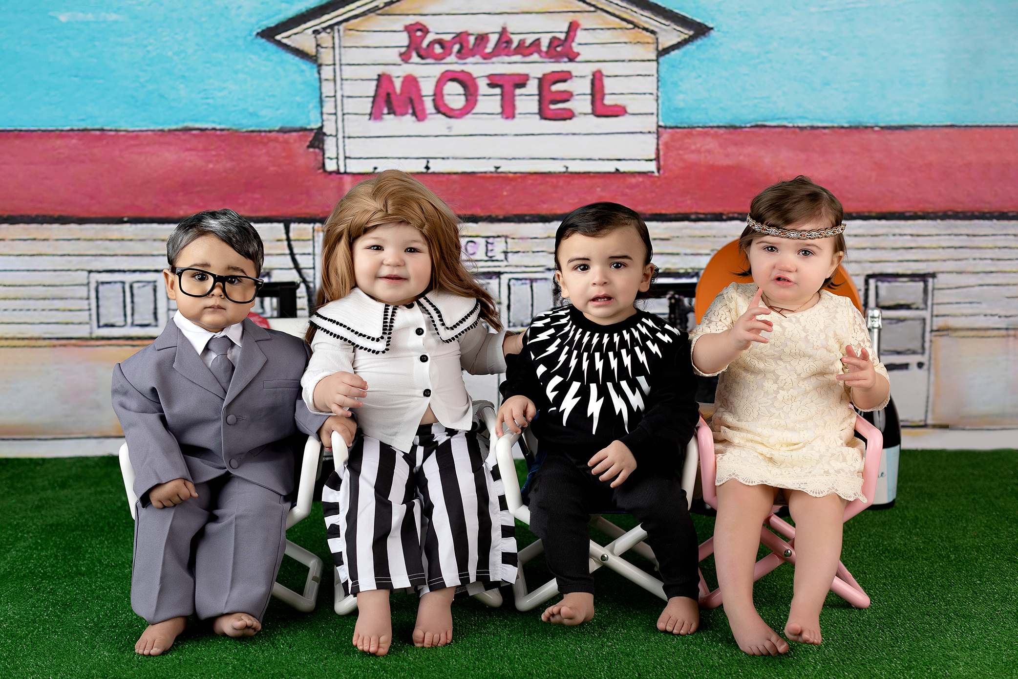 Four babies dressed in costumes sit in front of a backdrop with a Rosebud Motel sign, referencing the TV show Schitts Creek. Each baby is dressed as a different character from the show. Recreated by CT cake smash photographer.
