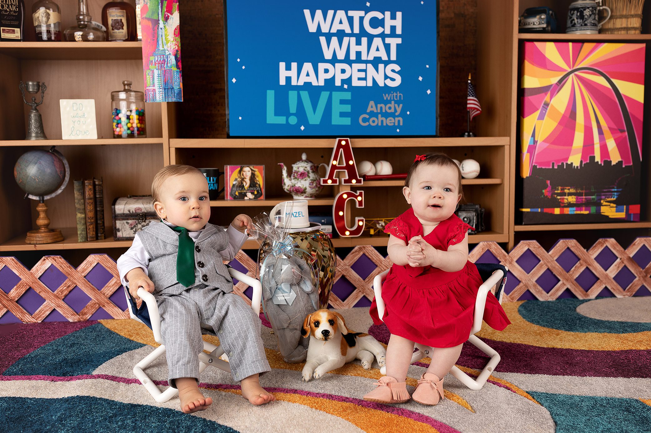 Two babies sit on small white chairs in a colorful room. The baby on the left wears a gray suit, and the baby on the right wears a red dress. A toy dog and large baby bottle are between them. A bright set and TV are behind them.