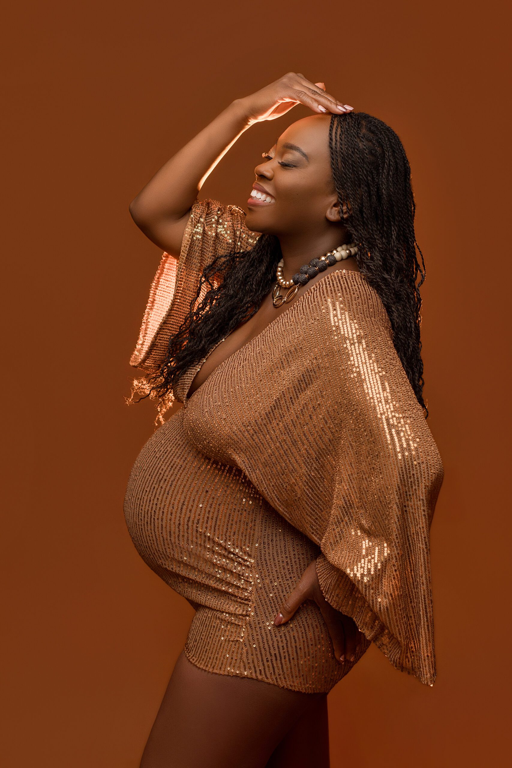 A smiling pregnant woman in a sparkly gold dress poses against a brown background, with one hand on her belly and the other hand resting on her head. She has long braided hair and wears a chunky necklace.