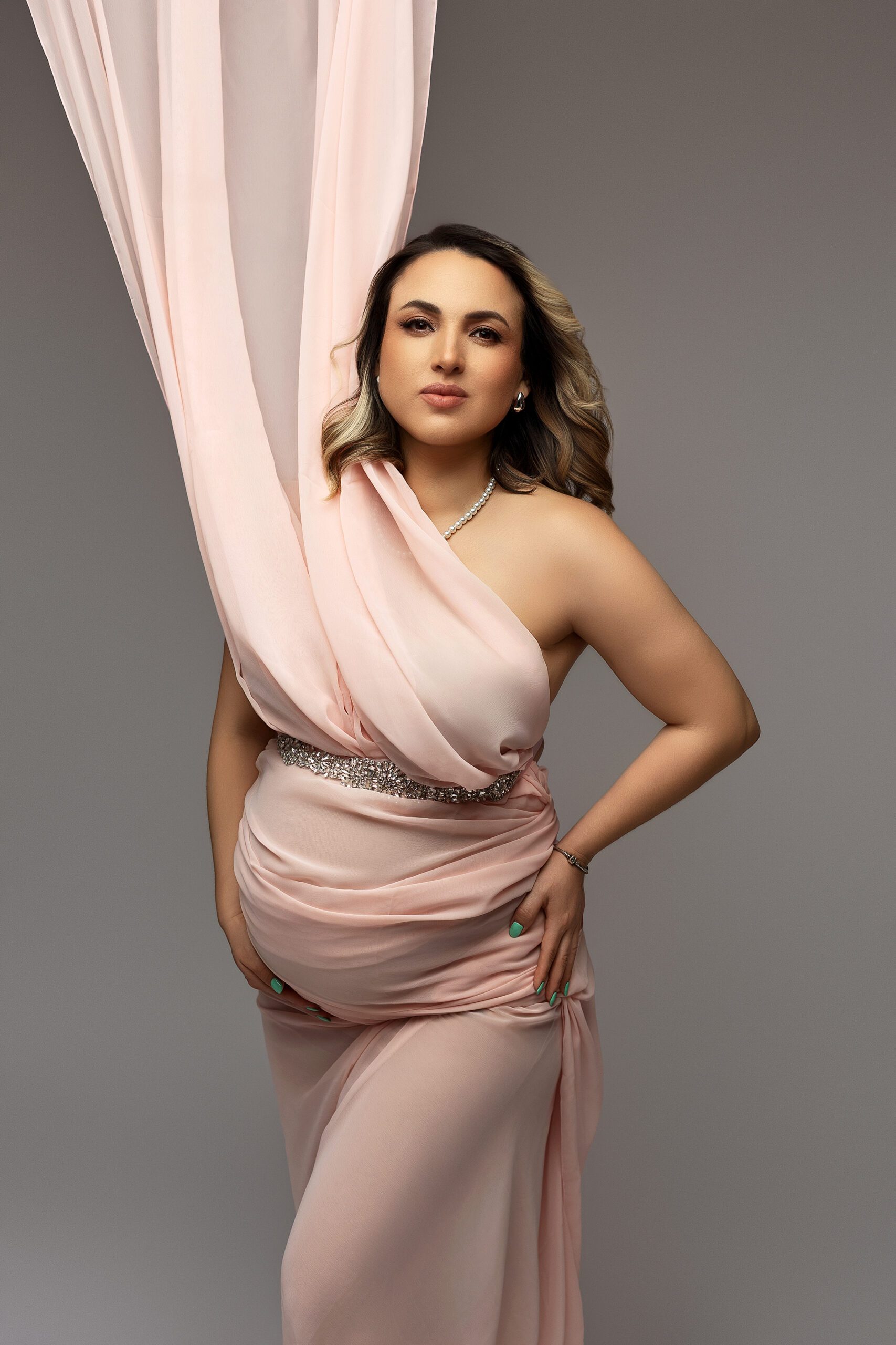 A pregnant woman poses confidently in a flowing, light pink dress with sheer fabric draped above her. She rests one hand on her belly and the other on her hip, standing against a neutral gray background during her CT maternity photos.