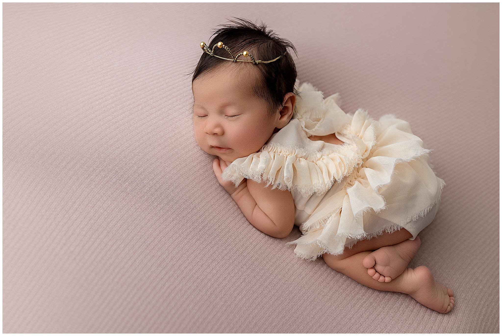 A sleeping baby lies on a soft pink surface, dressed in a frilly, cream-colored outfit. Capturing the essence of newborn photos, the baby has a delicate headband with tiny decorations, resting peacefully with eyes closed and arms gently tucked under.
