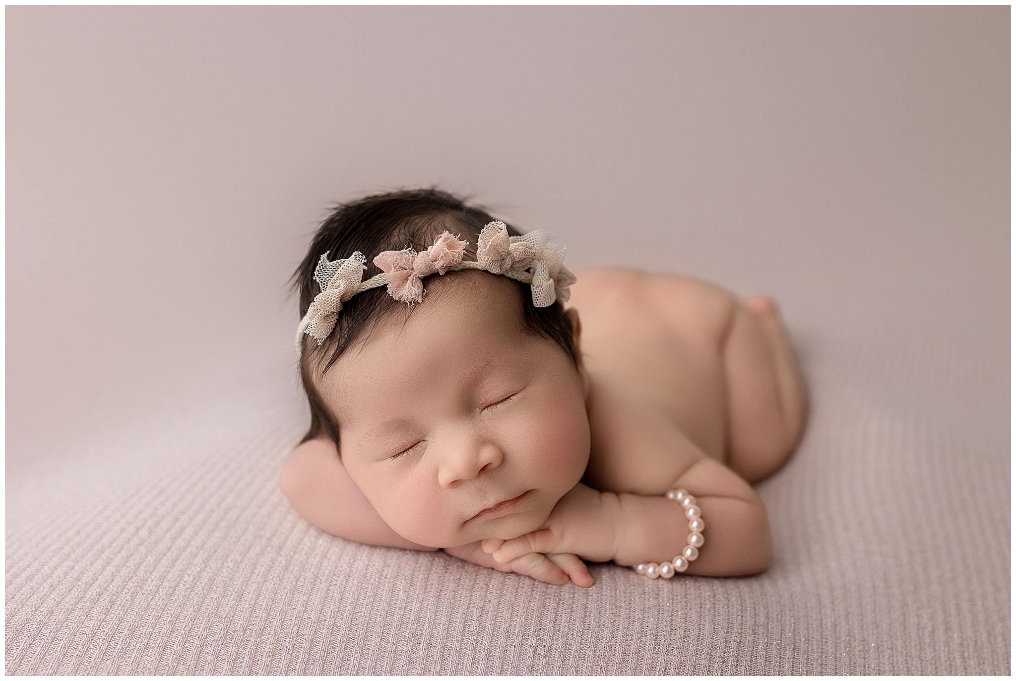 A sleeping newborn baby lies on a soft pink blanket. Their head rests on folded arms, adorned with a pink and white floral headband and a pearl bracelet on the wrist, creating a serene and delicate scene.