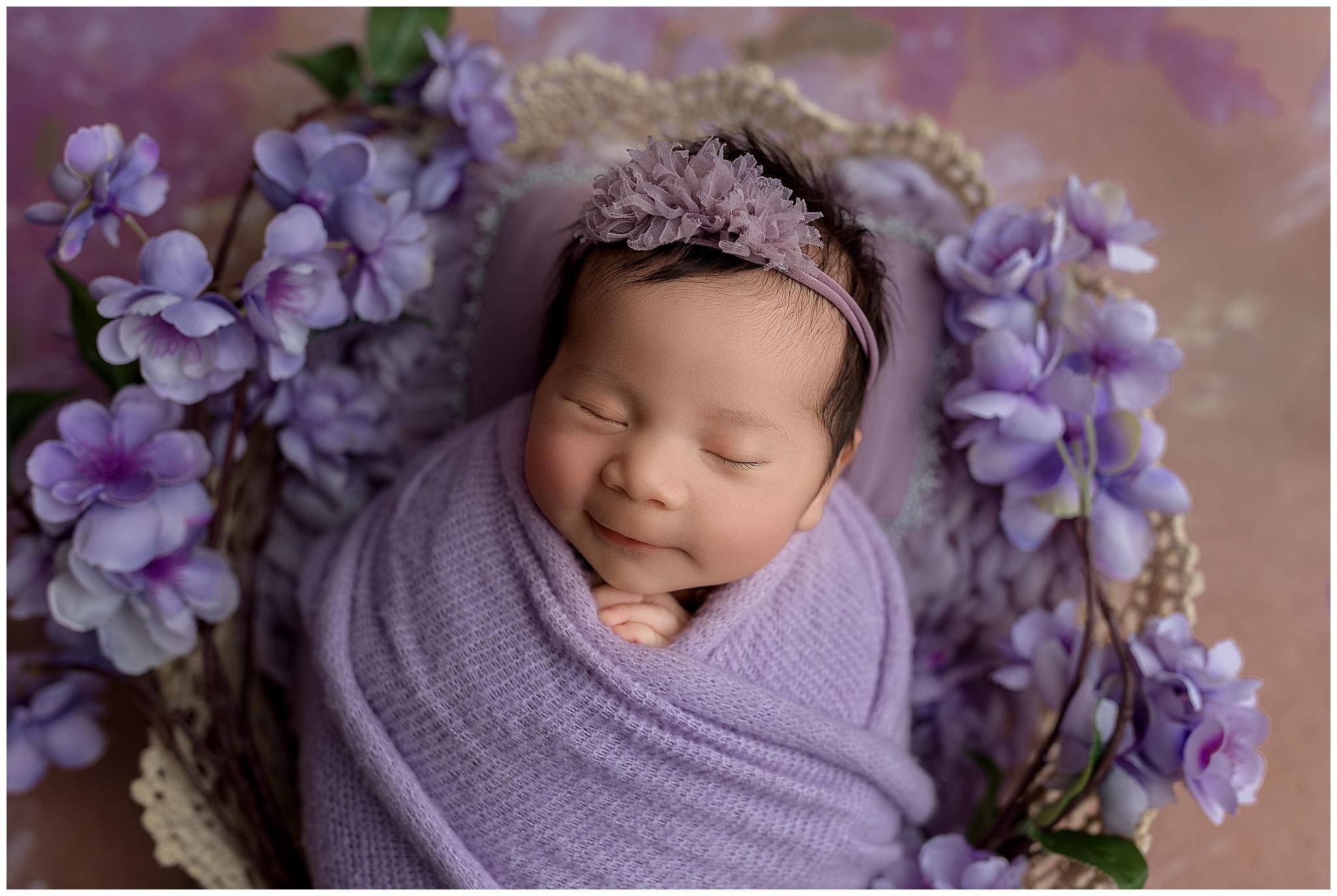 A newborn baby is peacefully sleeping, wrapped in a soft purple blanket. The baby is wearing a purple floral headband, surrounded by violet flowers, lying on a textured, neutral-toned surface. The scene conveys calmness and innocence.