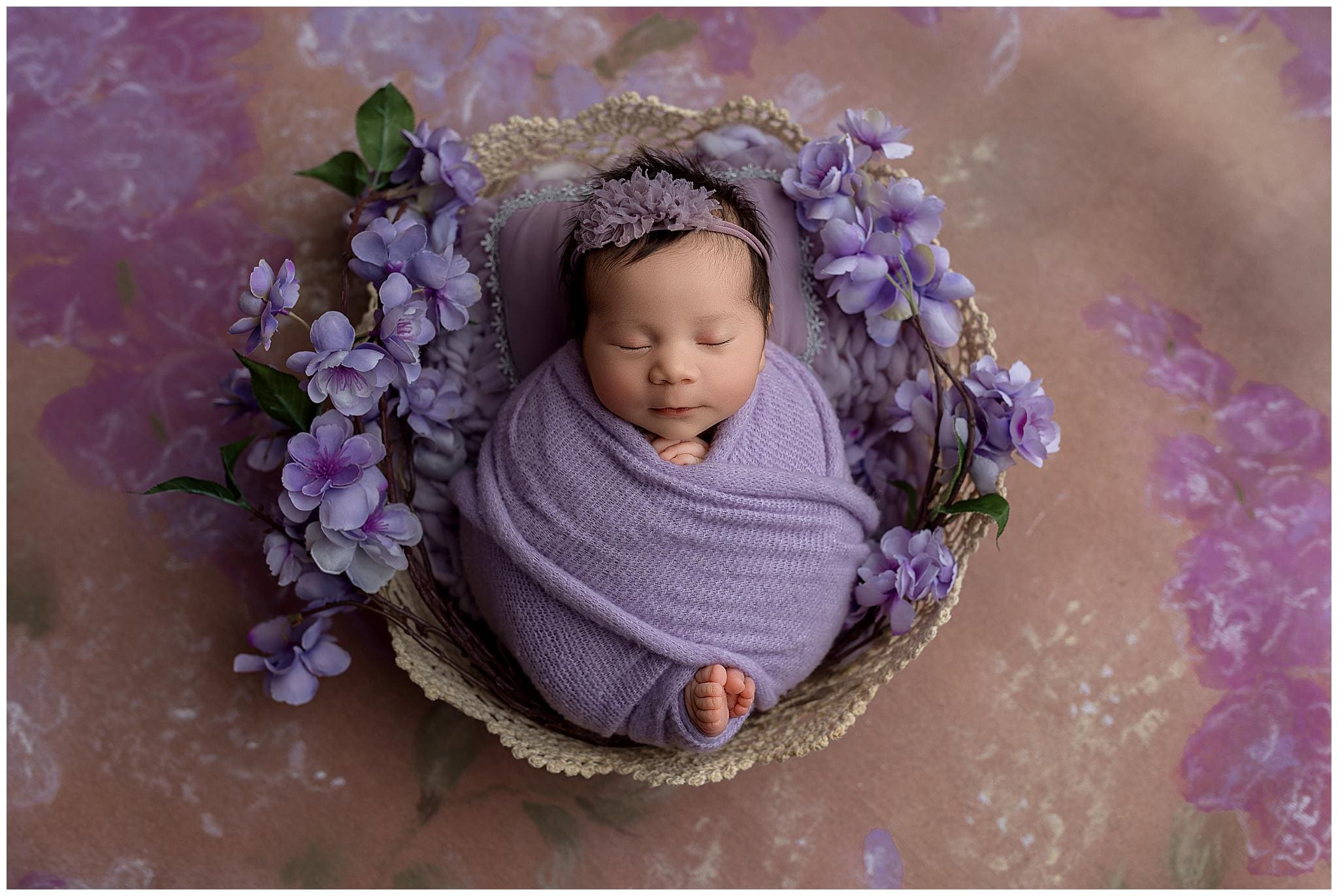 A newborn baby peacefully sleeps wrapped in a purple blanket, nestled in a decorative basket. The setup is adorned with purple flowers and set against a soft pink and purple floral backdrop. The baby wears a purple headband.