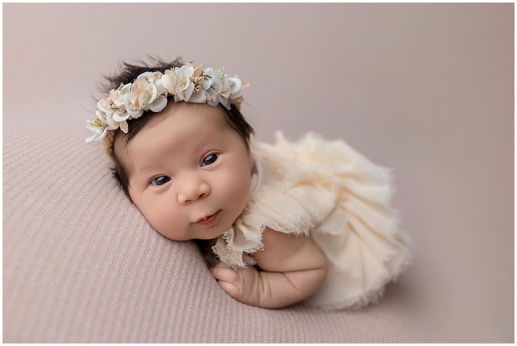 A baby wearing a cream-colored outfit with ruffled sleeves and a floral headband lies on a soft, textured surface. Captured by a talented newborn photographer in CT, the baby gazes directly at the camera with a gentle expression.
