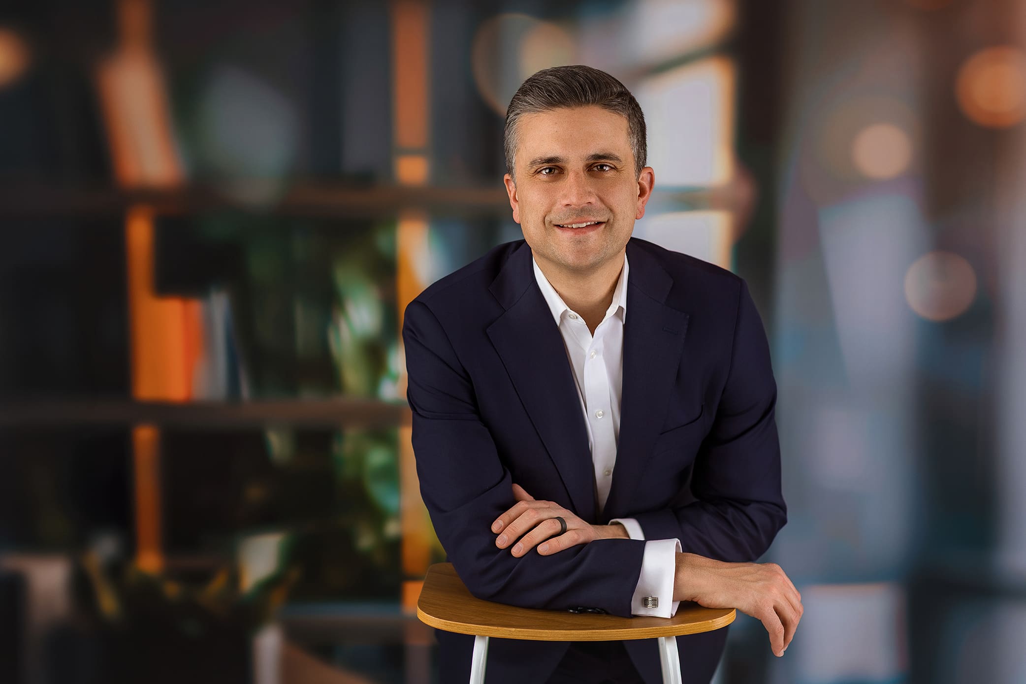 A man in a dark suit and white shirt is seated, leaning forward with his arms on a wooden stool. He is smiling, with a blurred background of warm colors and circles of light resembling bokeh used for his professional headshot in Connecticut.