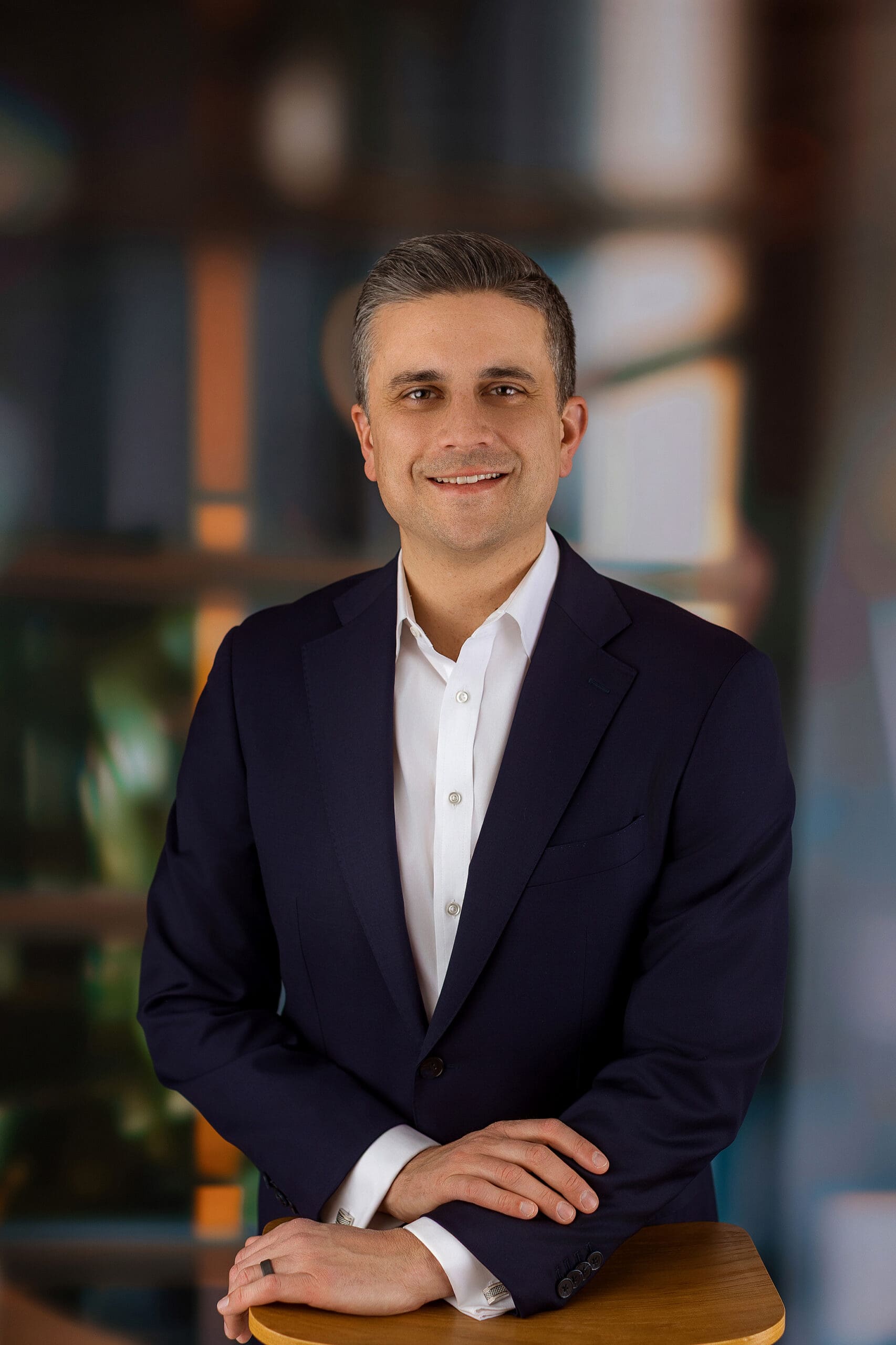 A smiling man in a dark suit and white shirt poses confidently, standing with hands resting on a wooden chair. The background is blurred with warm and cool tones, suggesting a professional setting for his Connecticut headshot.