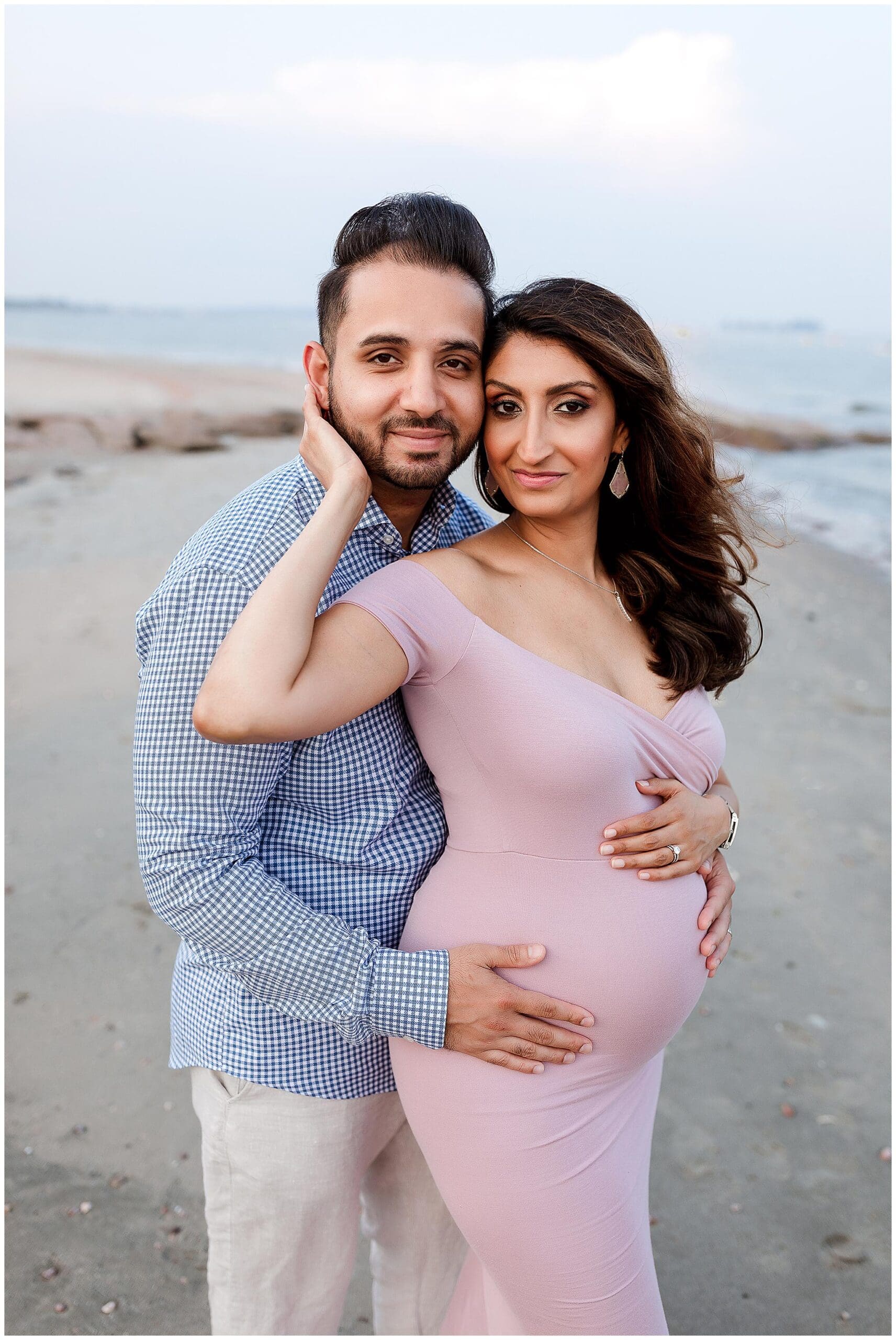 A couple poses on a beach, with the man in a blue checkered shirt and white pants, and the woman in a light pink dress, cradling her pregnant belly. The ocean and sky are in the background.