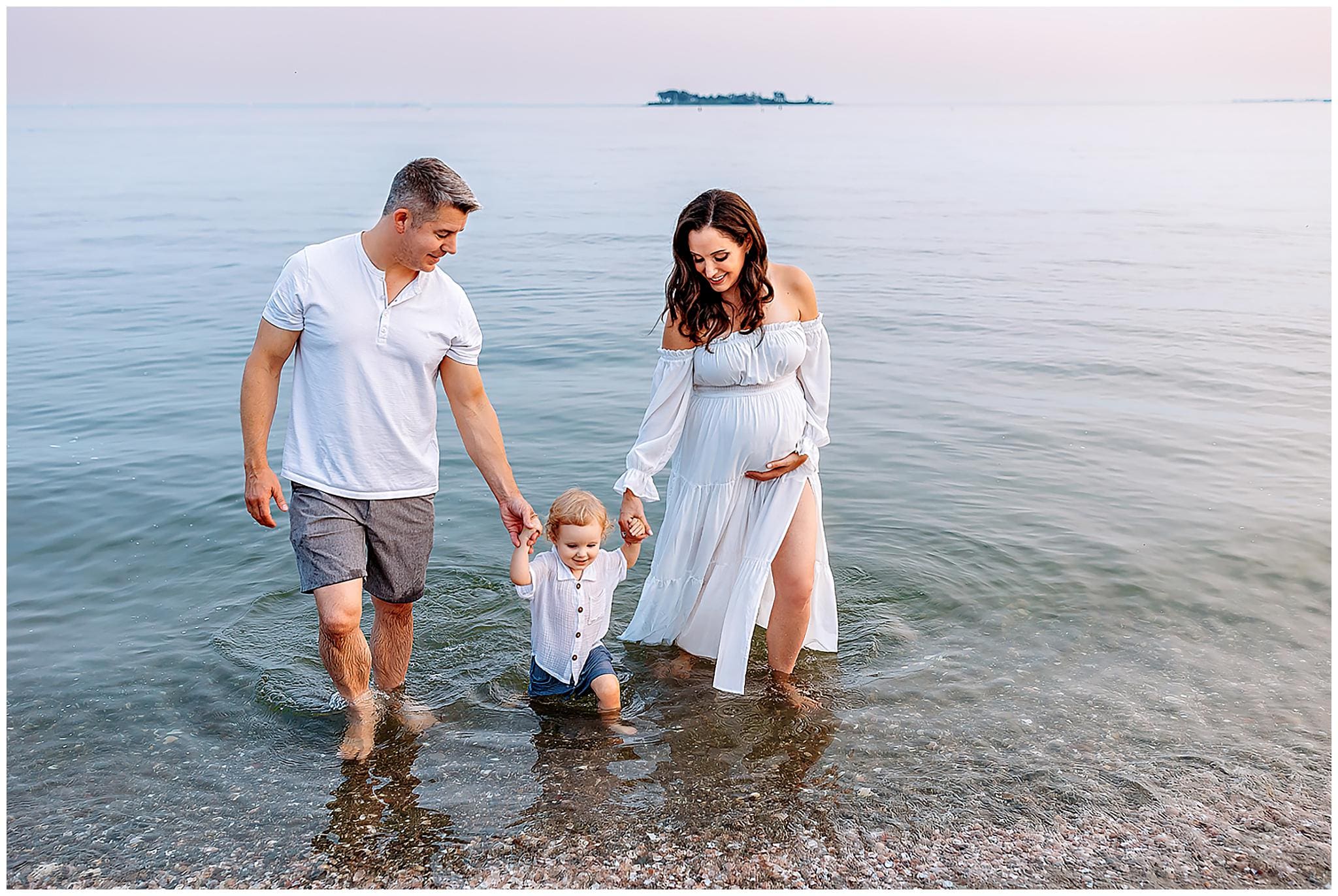 A family of three strolls along the beach, captured beautifully by a Connecticut maternity photographer. The father and pregnant mother hold hands with their young child, who splashes in the shallow water. Their smiles reflect the joy beneath a clear sky, as the ocean stretches into the horizon.