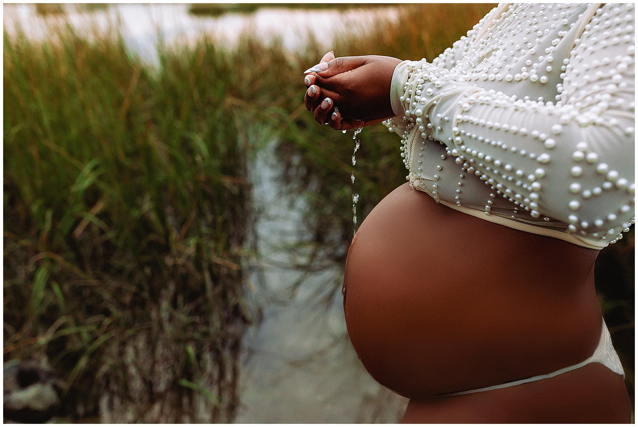 During a maternity photo session, a pregnant person holds water in their cupped hands near a marsh, with grass gently swaying in the background. They wear a white, pearl-embellished top, highlighting their beautifully rounded belly.