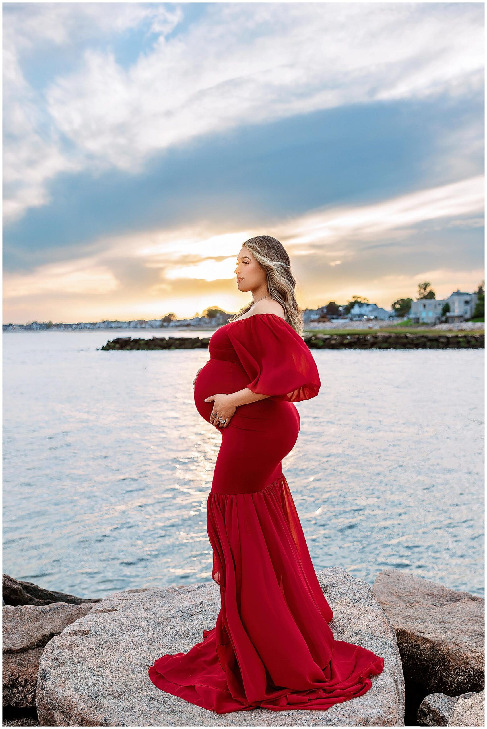 A pregnant woman in a flowing red dress stands on rocks by a calm beach in CT waterfront during sunset. Her hand gently cradles her belly, and a soft, cloudy sky fills the background.
