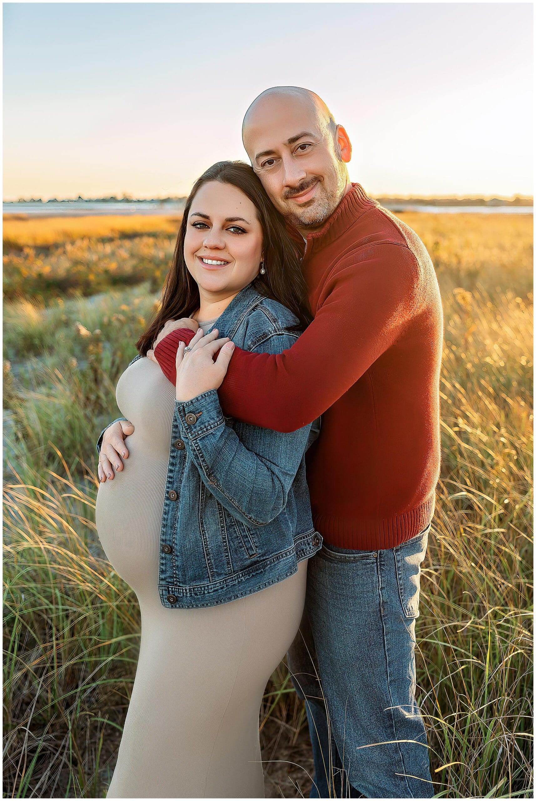 A couple stands in a field during sunset. The woman is pregnant, wearing a beige dress and denim jacket, and the man is wearing a red sweater and jeans. He stands behind her, gently embracing her. They both smile at the camera and their Connecticut Maternity Photographer.
