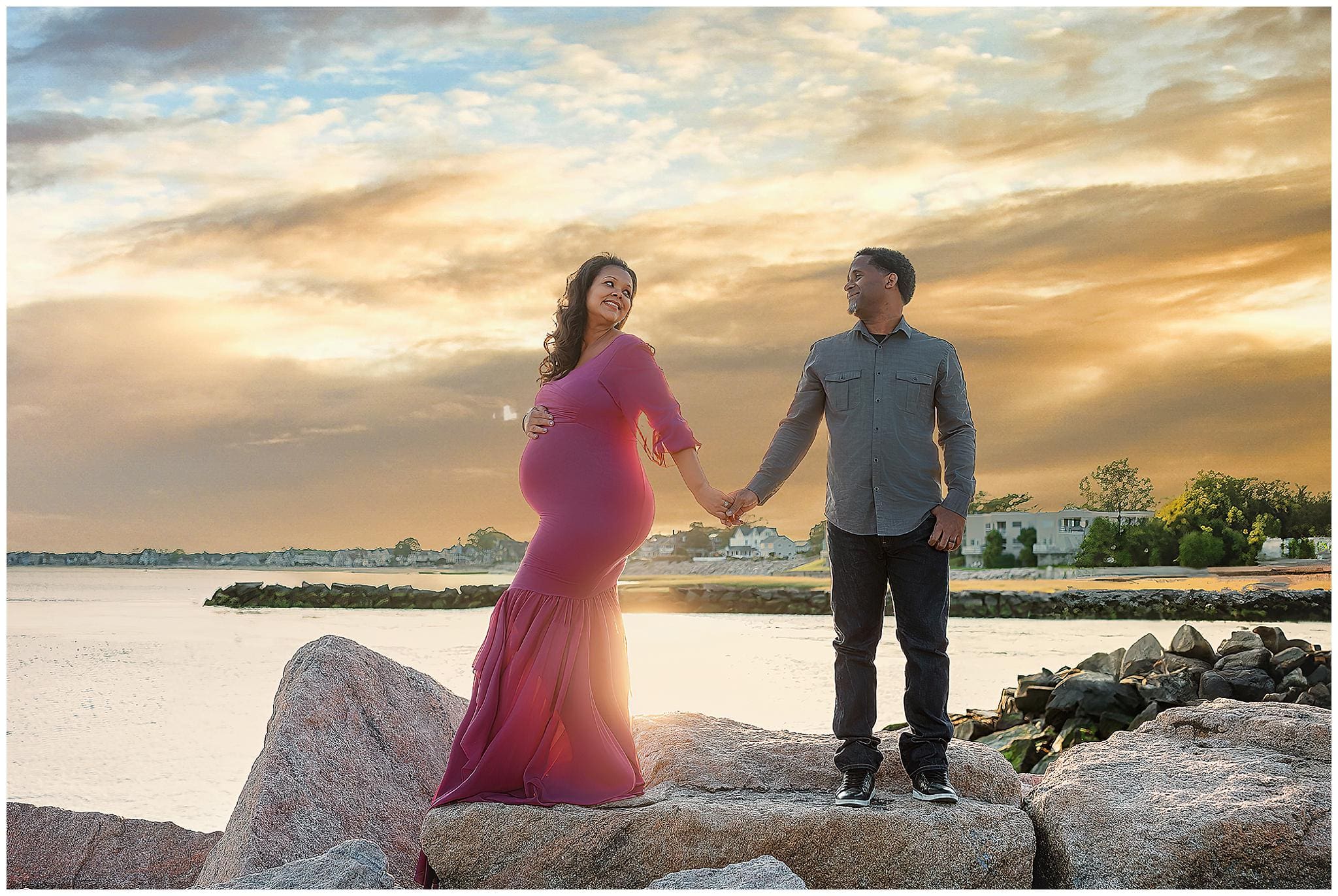 A pregnant woman in a flowing purple dress stands on rocks by the water, smiling at a man holding her hand. He is wearing a gray shirt and jeans. The sky is golden during sunset, and distant houses and trees are visible.