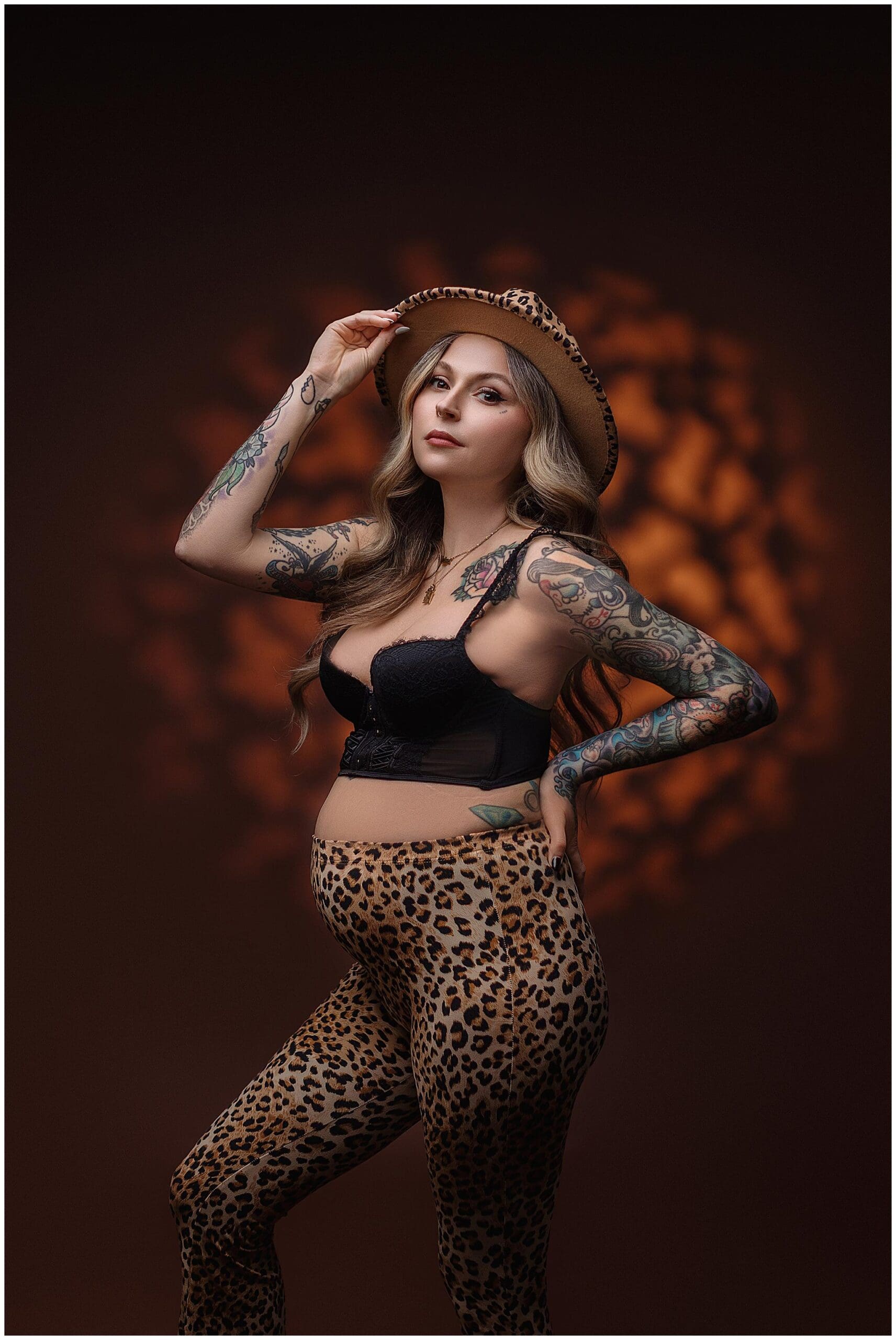 Pregnant woman in Connecticut with long hair poses confidently wearing a leopard-print hat and pants, paired with a black top. She displays tattoos on her arms, standing against a brown background with abstract lighting.