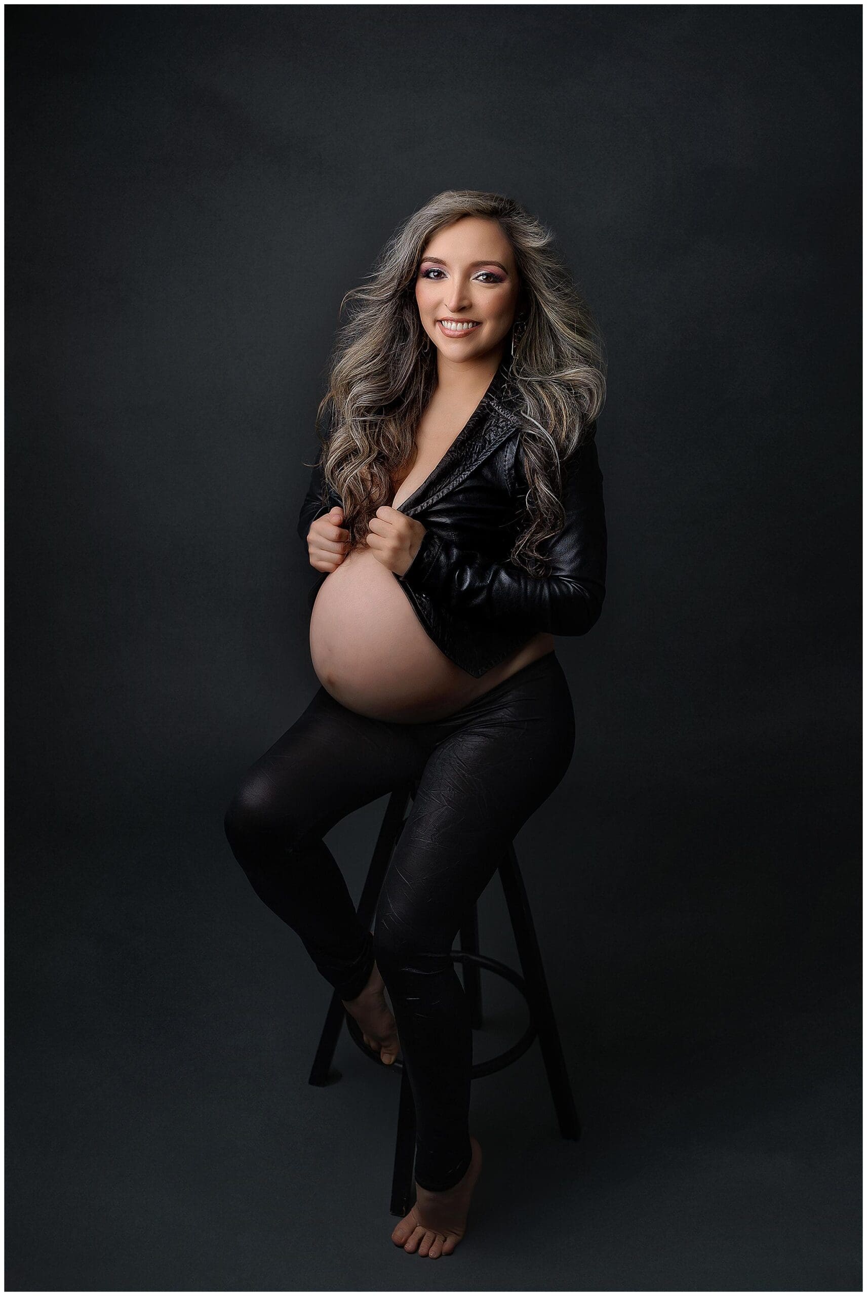 A pregnant woman with long, wavy hair sits on a stool against a dark background. Shes wearing a black leather jacket partially open to reveal her belly, and black pants. She is barefoot and smiling at the camera.