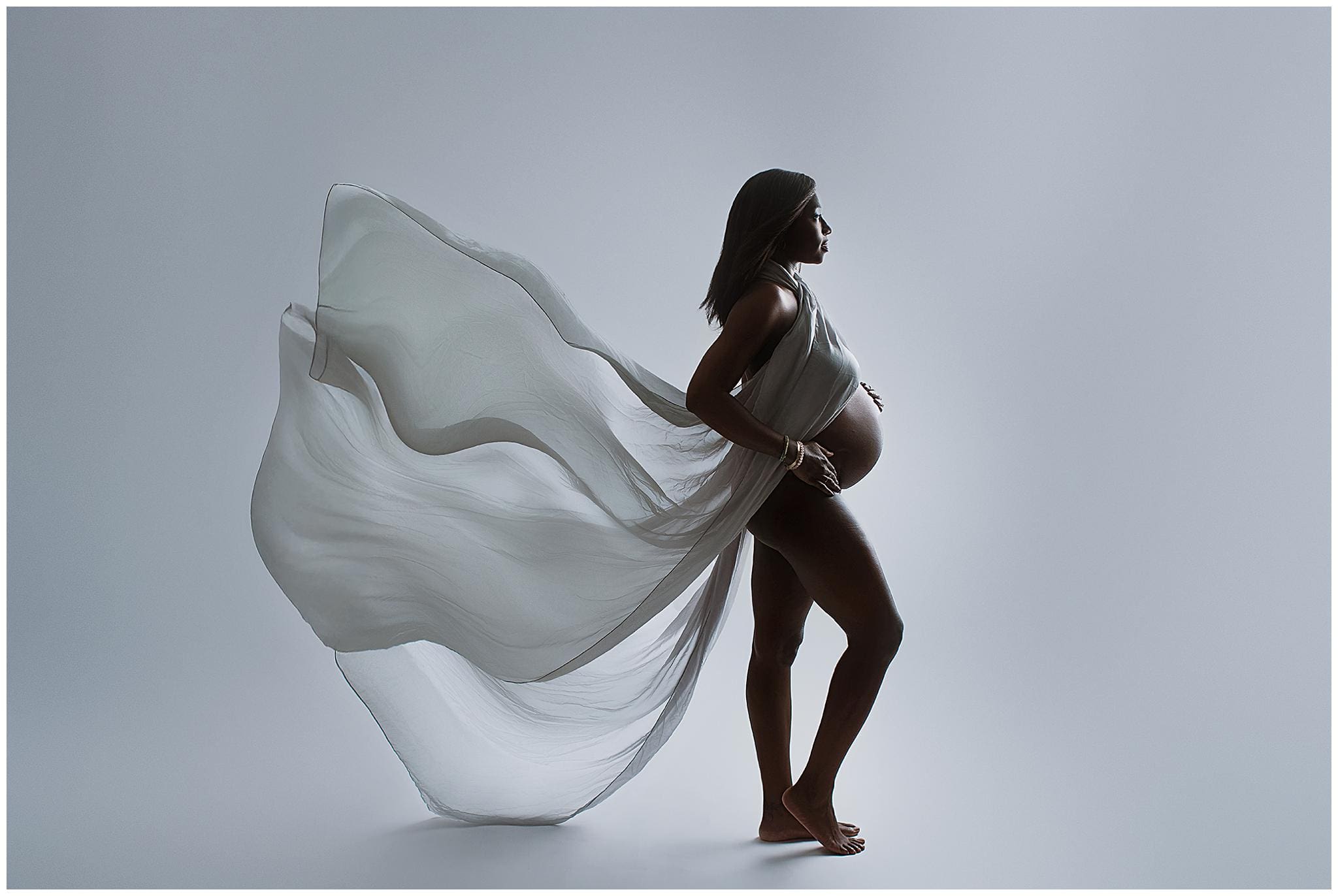 A pregnant woman stands in profile against a light background, embodying the essence of maternity photos. Her flowing translucent dress billows behind her as she tenderly holds her belly, exuding a serene and graceful presence.