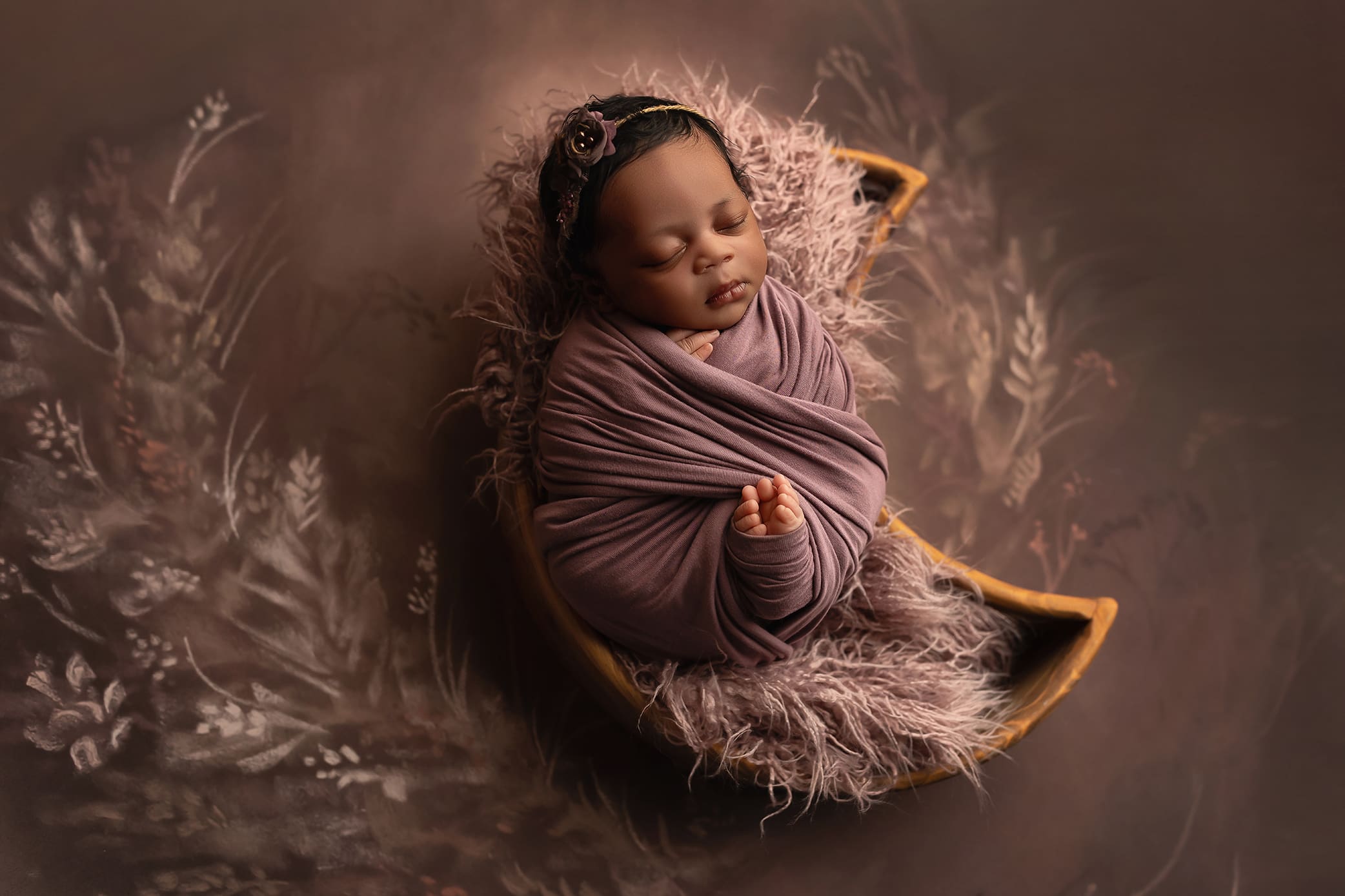 A sleeping newborn wrapped in a mauve blanket rests on a furry cushion in a crescent moon-shaped basket, surrounded by soft, floral-patterned décor in warm brown tones posed by a top Connecticut newborn photographer.