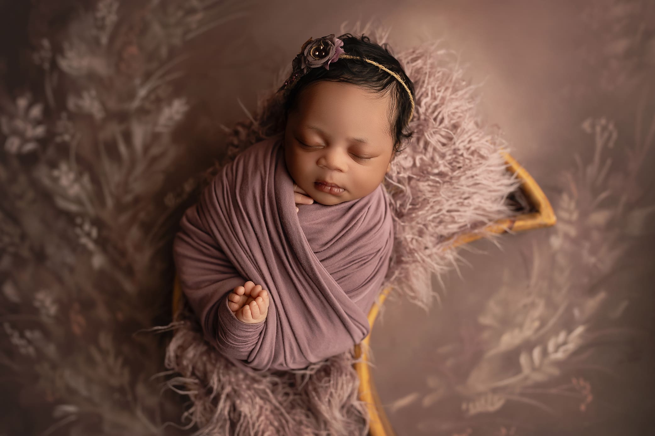 A newborn baby sleeps peacefully, swaddled in a mauve wrap and lying on a fluffy blanket. The baby wears a delicate floral headband, and the background is soft with floral patterns in muted tones perfectly photographed by Connecticuts favorite newborn photographer.