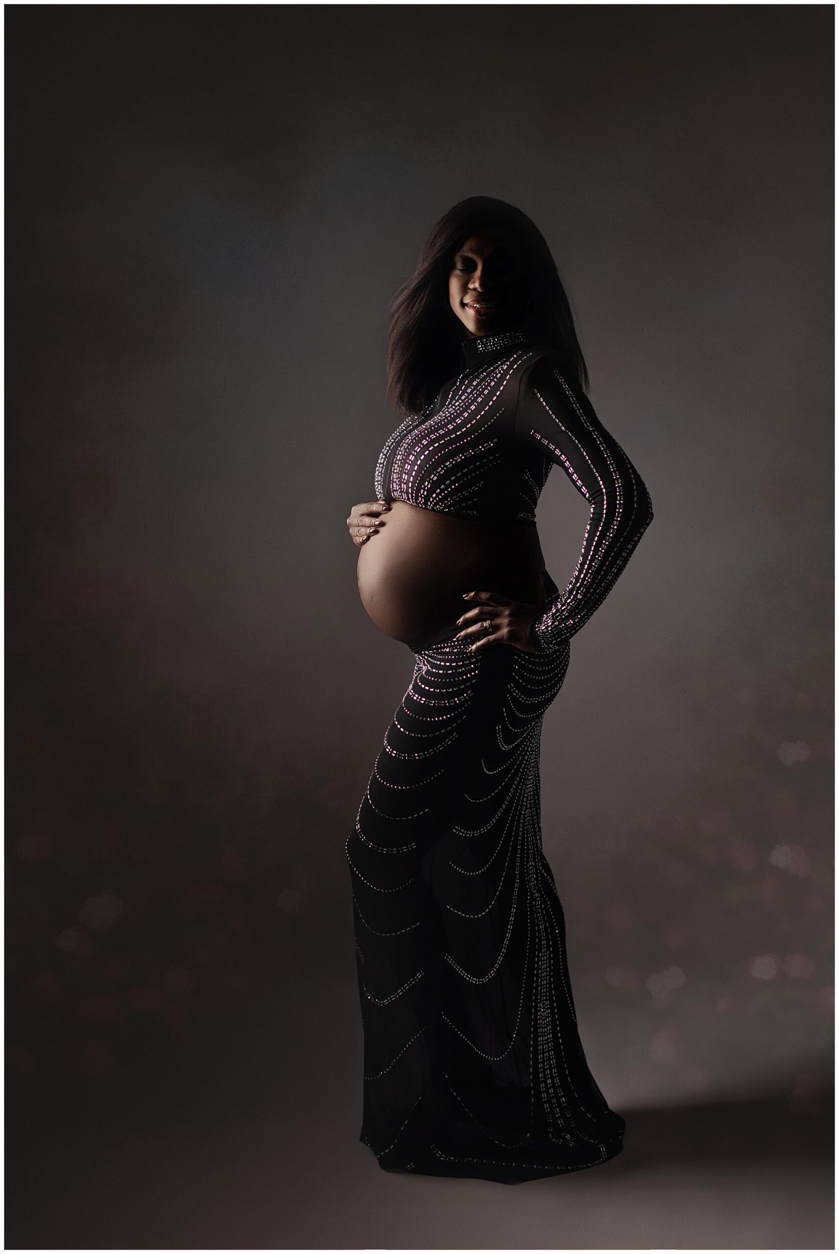 Pregnant woman in a long, fitted black dress with sparkling details, holding her belly. She stands against a dark, softly lit background, with a serene expression.