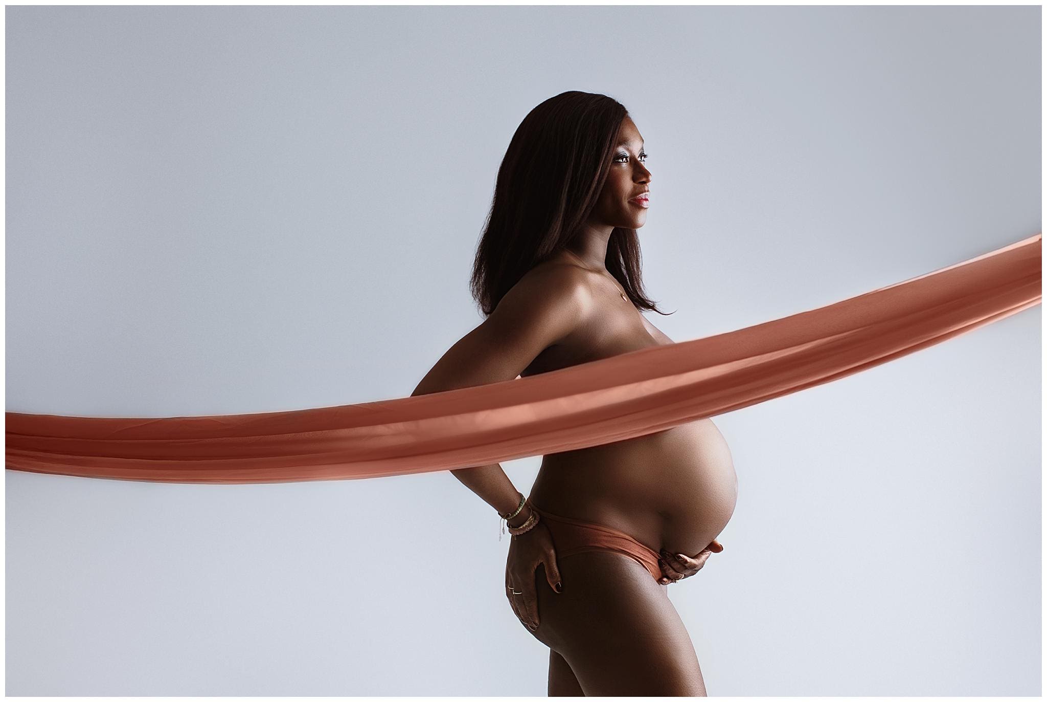 Pregnant woman poses elegantly, draped with a flowing sheer orange fabric. She stands in profile against a soft gray background, highlighting her baby bump and serene expression.