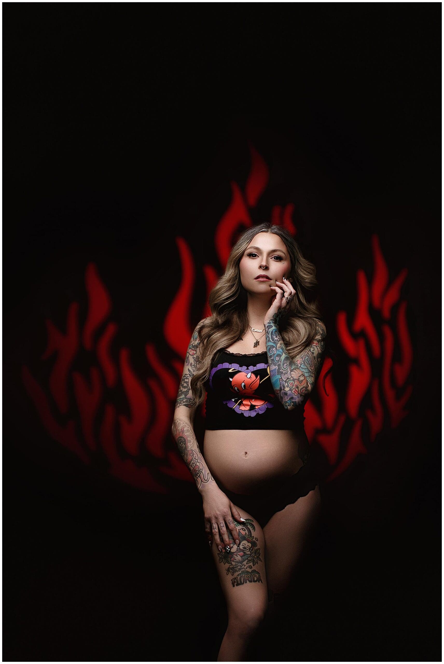 A pregnant woman poses confidently against a dark background with a red flame design during her maternity photos. She has long hair, tattoos covering her arms and legs, and is wearing a black crop top and underwear. She gazes at the camera with one hand on her face.