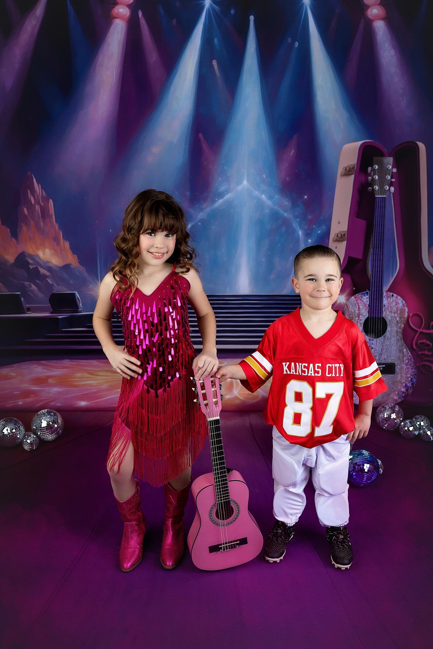 A girl in a shiny pink dress and a boy in a Kansas City jersey stand on stage under bright spotlights. A pink guitar sits between them amid the rock and roll backdrop, captured perfectly by a Connecticut child photographer known for vibrant storytelling through imagery.