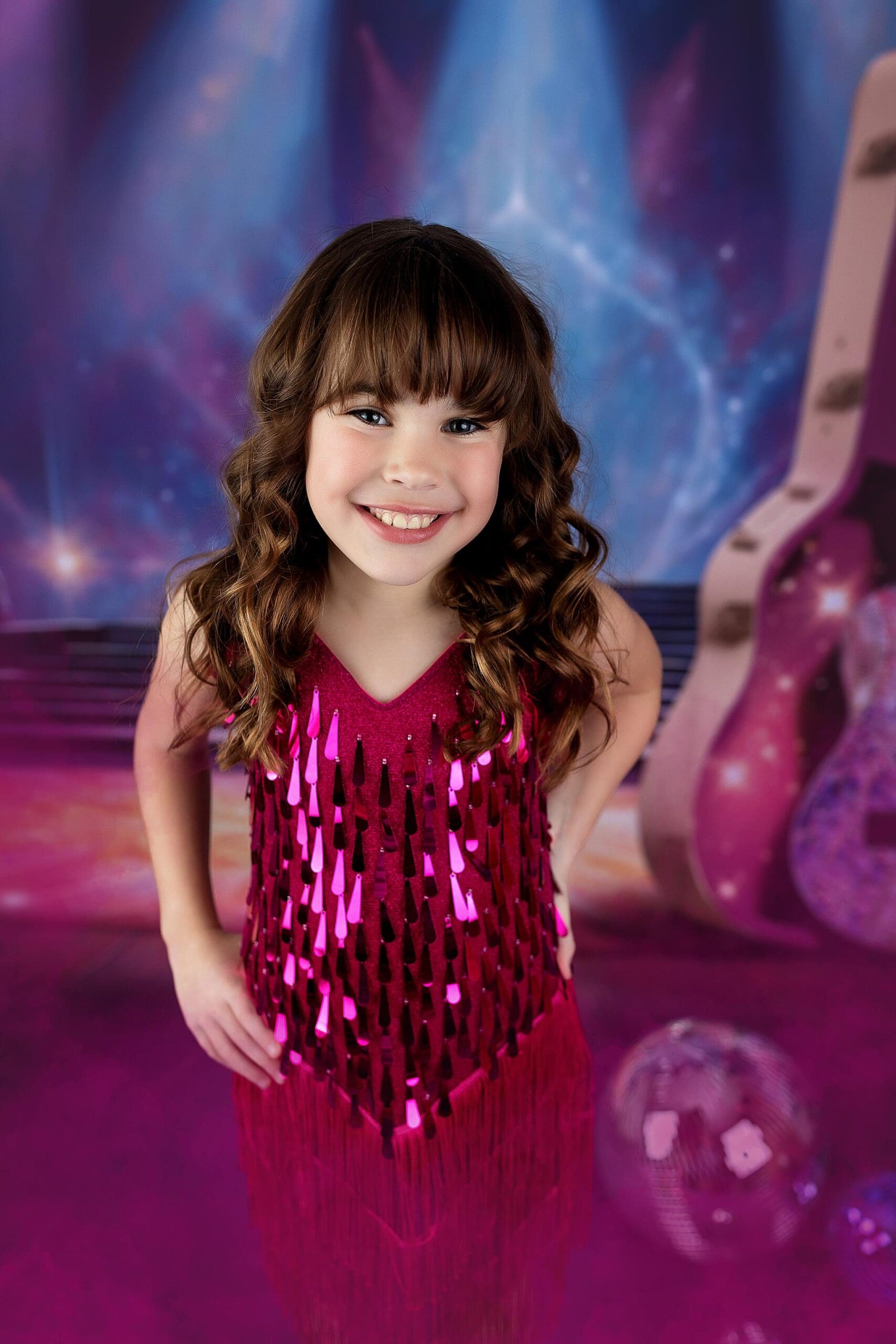 A smiling young girl with long, wavy brown hair stands confidently in a sparkly pink dress with fringe, capturing the essence of child photography. The background features a stage with vibrant blue and purple lighting and a large acoustic guitar.