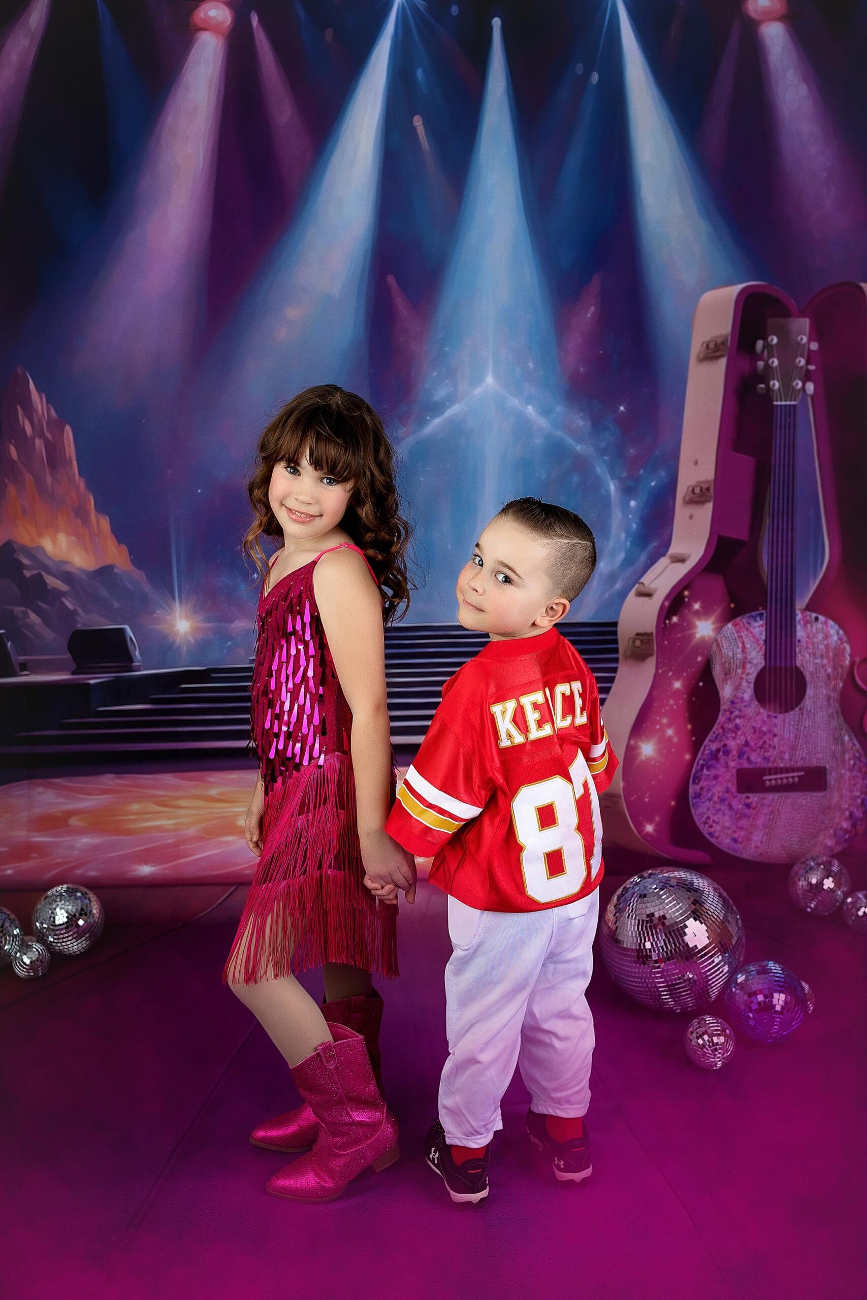 Two children stand holding hands on a stage with colorful lights. The girl wears a sparkly pink dress and boots, while the boy sports a red jersey and white pants. Disco balls and a guitar are in the background, creating a vibrant, musical atmosphere in a studio in Connecticut.