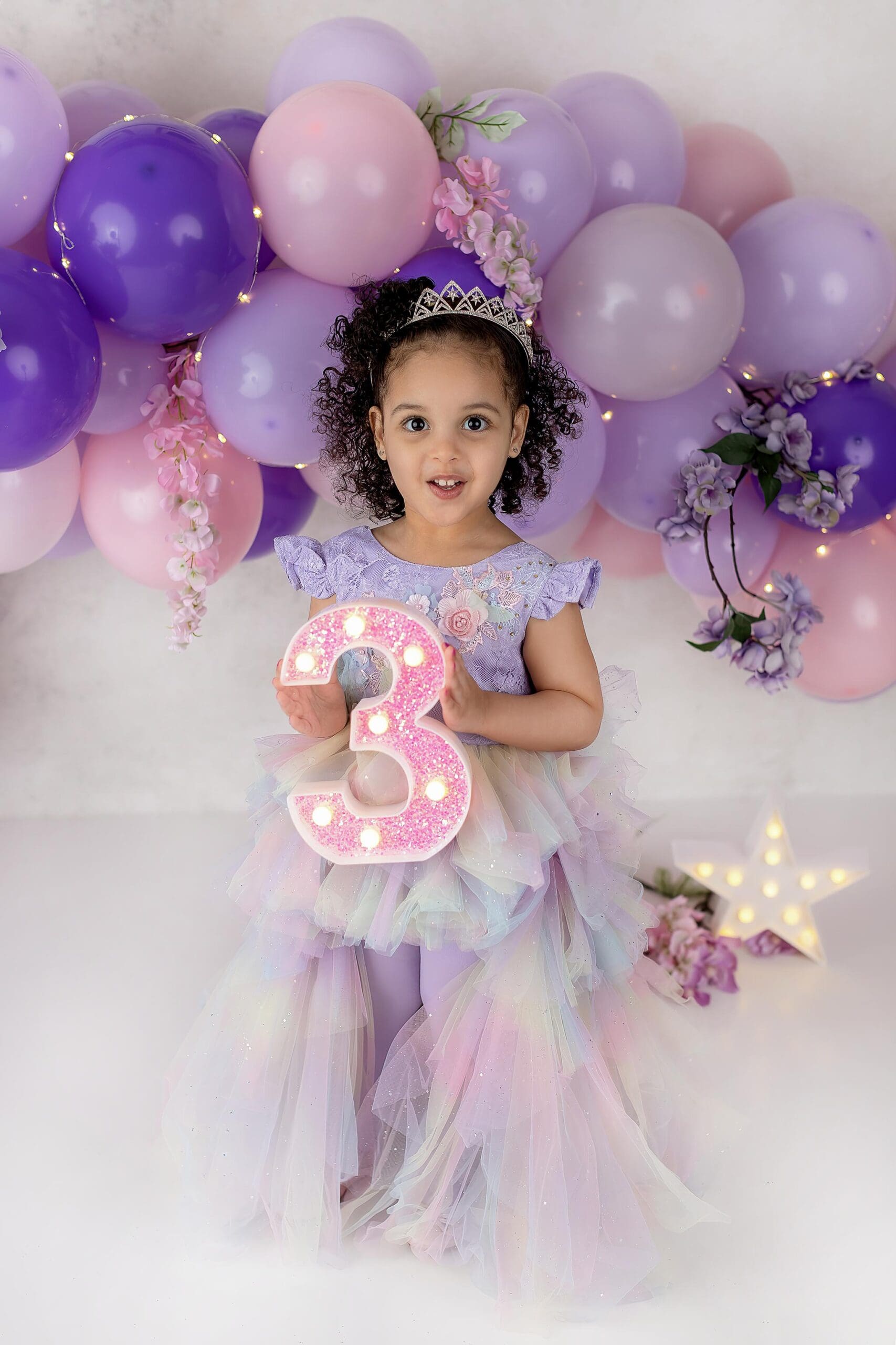 A young girl in a pastel rainbow dress holds a light-up number three. Shes wearing a tiara, standing in front of a backdrop with purple and pink balloons and flowers. Captured in Connecticut, this childs enchanting moment is completed by a star-shaped light on the floor.