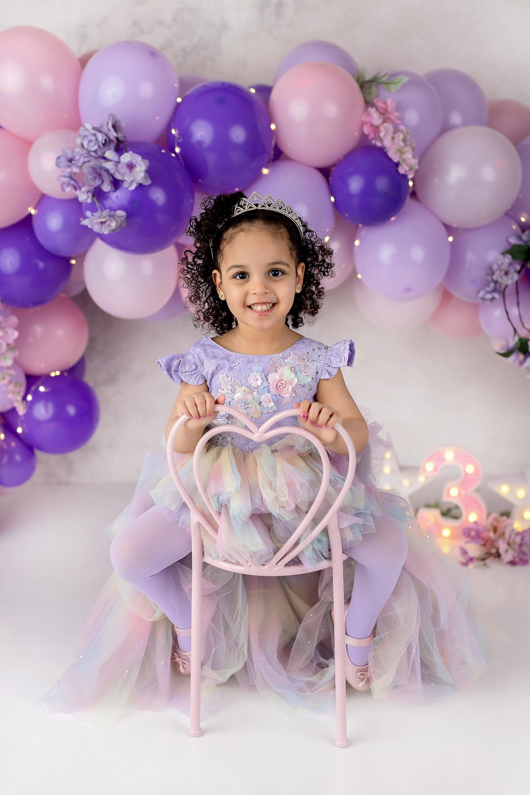 A young girl in a colorful dress and tiara sits on a pink heart-shaped chair, embodying the joy captured through CT children photography. She smiles in front of a backdrop of pink, purple, and lavender balloons, with a glowing number three on the floor beside her.