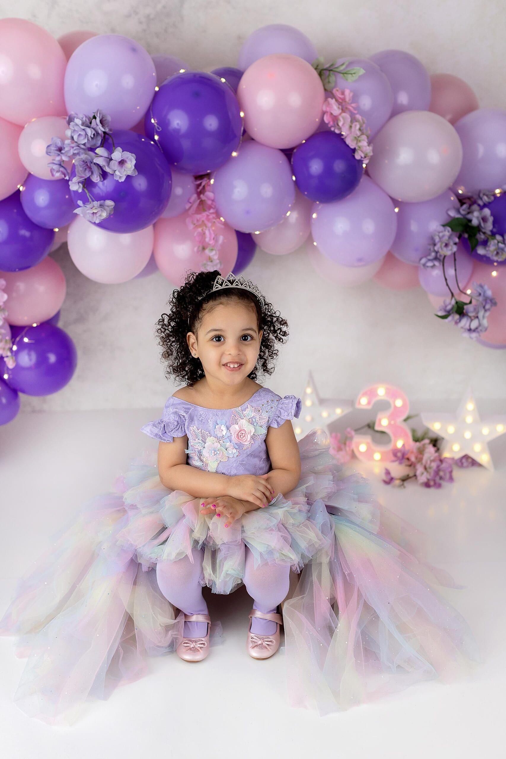 A young girl beams in front of purple and pink balloons, captured beautifully by a talented CT child photographer. Shes wearing a lavender dress with a colorful tulle skirt and a tiara. A lit number 3 and star decorations adorn the floor beside her, completing the festive scene.