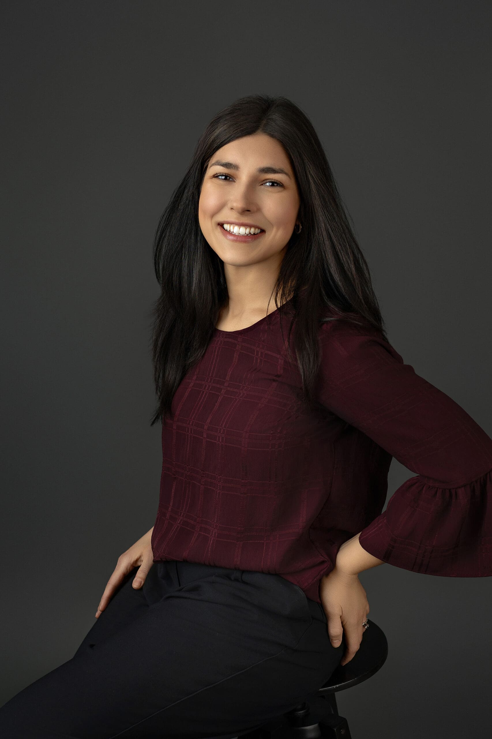 A woman with long dark hair, wearing a maroon blouse and black pants, is sitting on a stool against a dark gray background, smiling with her hands resting on her hips during her studio branding session in Connecticut.