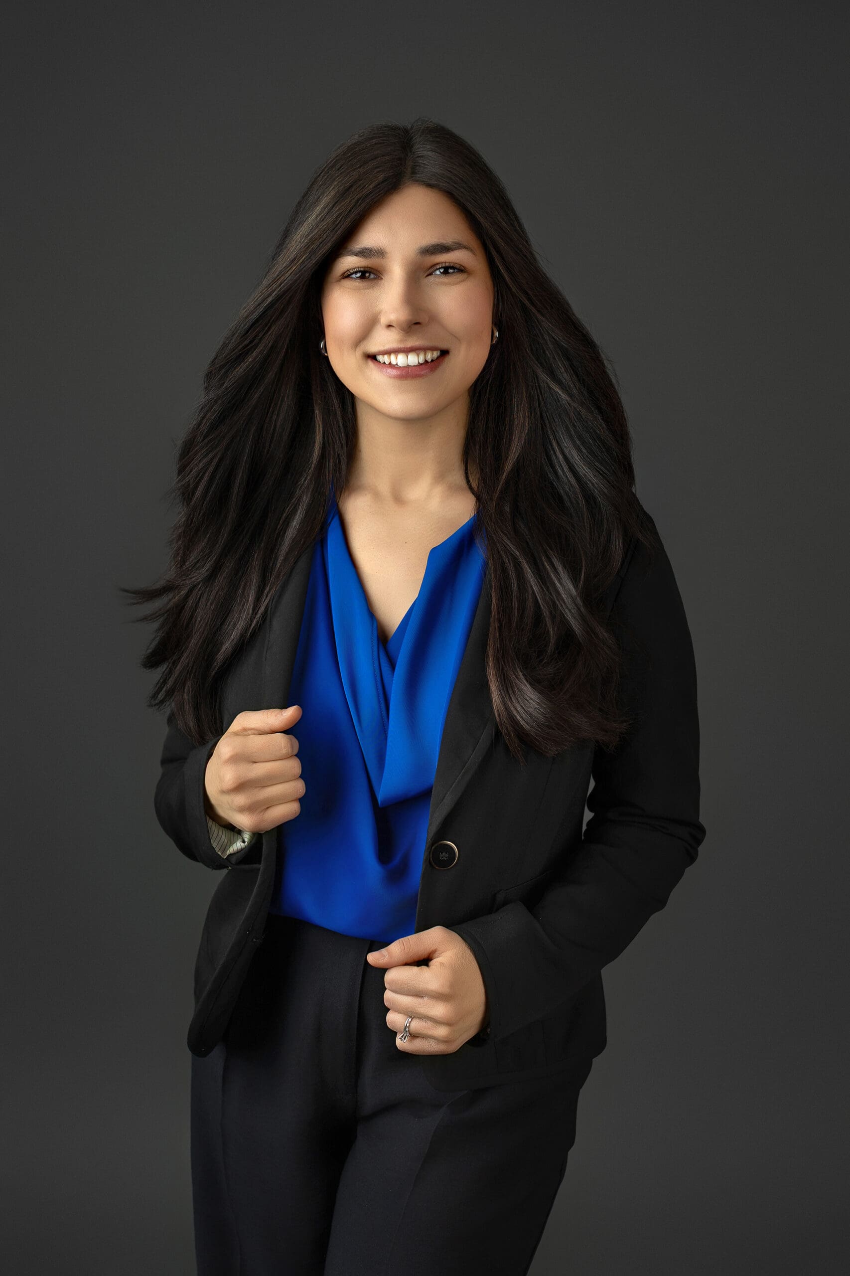 A woman with long dark hair smiles, wearing a black blazer over a blue blouse. She stands against a dark gray background, with one hand holding her blazer lapel and the other by her side.