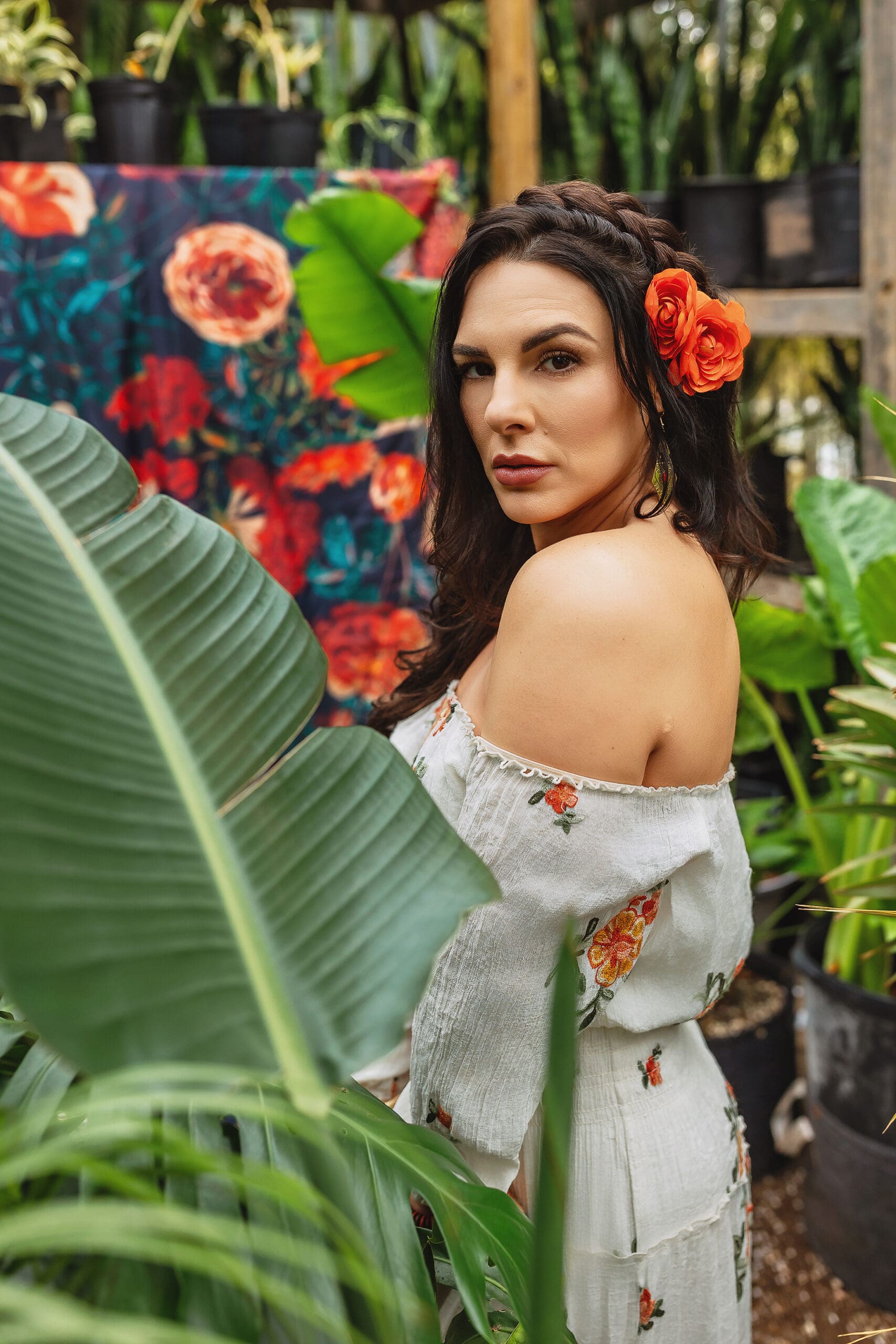 In a headshot photography session in CT, a woman with long dark hair adorned with a red-orange flower poses gracefully among lush green plants. She wears an off-the-shoulder floral dress against the backdrop of a vibrant tapestry and potted plants. Captured by Headshot Photographer in Connecticut.